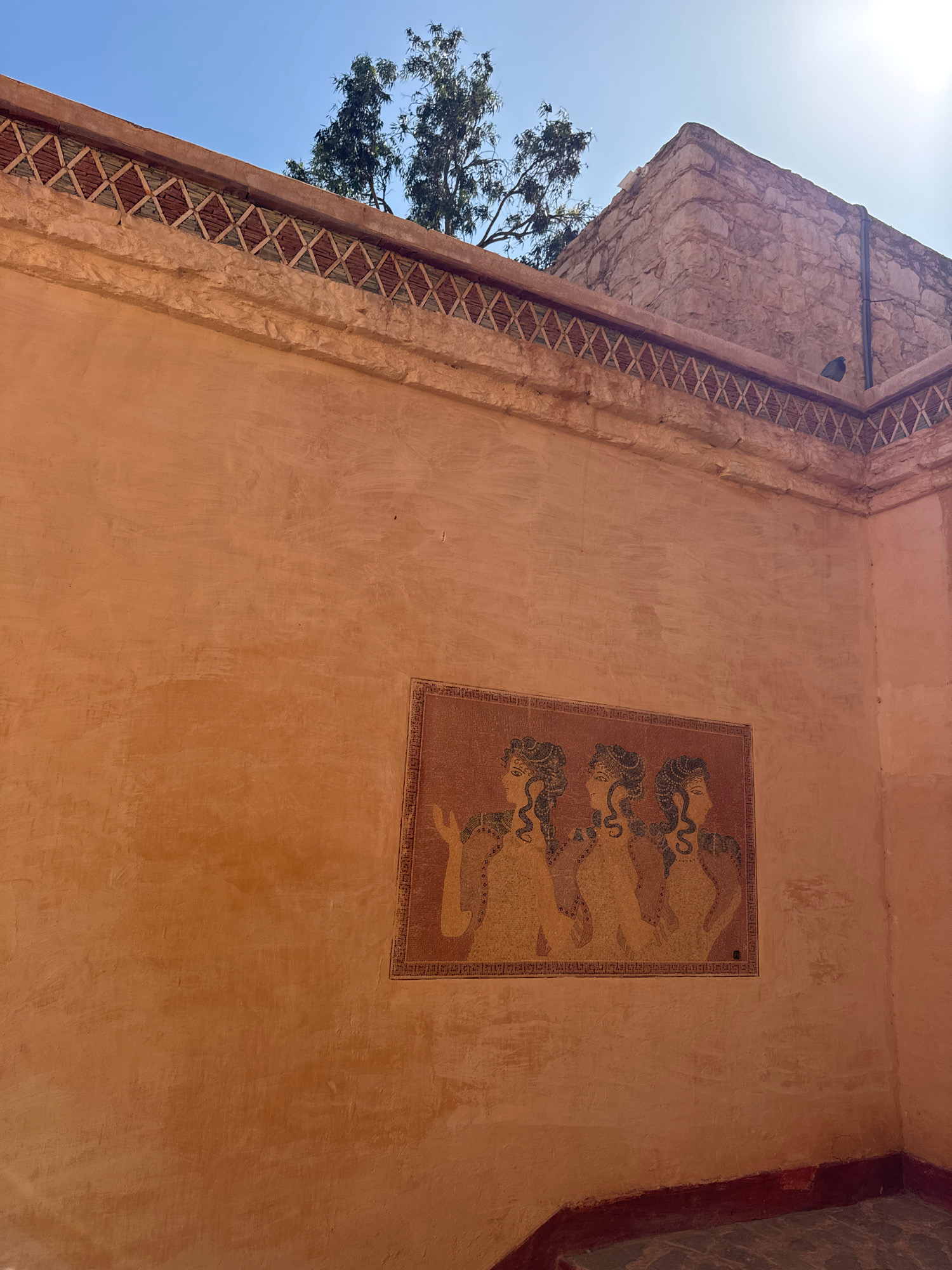The image depicts an interior courtyard or alcove with terracotta-colored walls. A mosaic artwork featuring three stylized female figures with serpentine hair is affixed to the wall. The mosaic is bordered by intricate patterns. Above the mosaic, along the upper edge of the wall, a decorative band of alternating reddish-brown and green triangular tiles is visible. In the background, a corner of a stone structure is present, along with the branches of a tree against a clear blue sky. The scene appears to be located within the walls of the Kasbah of Agadir Oufella, a historical fortress overlooking the city of Agadir, Morocco.
