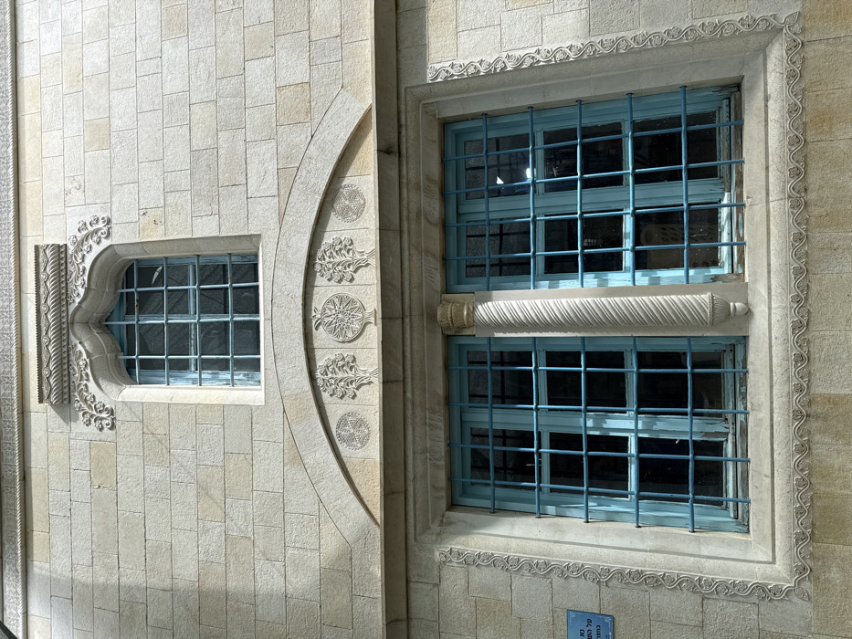 The image displays the exterior facade of a stone building, identified as being in Safed, Israel. The structure is constructed from light-colored, rectangular cut stone blocks. Two distinct levels of windows are visible. The upper level features a single, smaller arched window set within an ornate, carved stone frame with scrollwork. This window has a light blue frame and a rectangular metal grille. Below this, a wider, arched stone panel extends across the facade, featuring four carved circular motifs: two stylized trees flanking a central star-like design, and a geometric pattern on the far left. Directly beneath this panel are two rectangular windows, each with light blue frames and prominent metal grilles. These lower windows are separated by a carved, spiraling stone column. The frames of these lower windows are further adorned with vertical carved scrollwork borders and a wavy, leaf-patterned design along the bottom stone sill. A small blue sign with white Hebrew text is partially visible on the wall to the left of the lower windows. The building is illuminated by direct sunlight, highlighting the stone textures and carved details. No individuals are present in the frame.