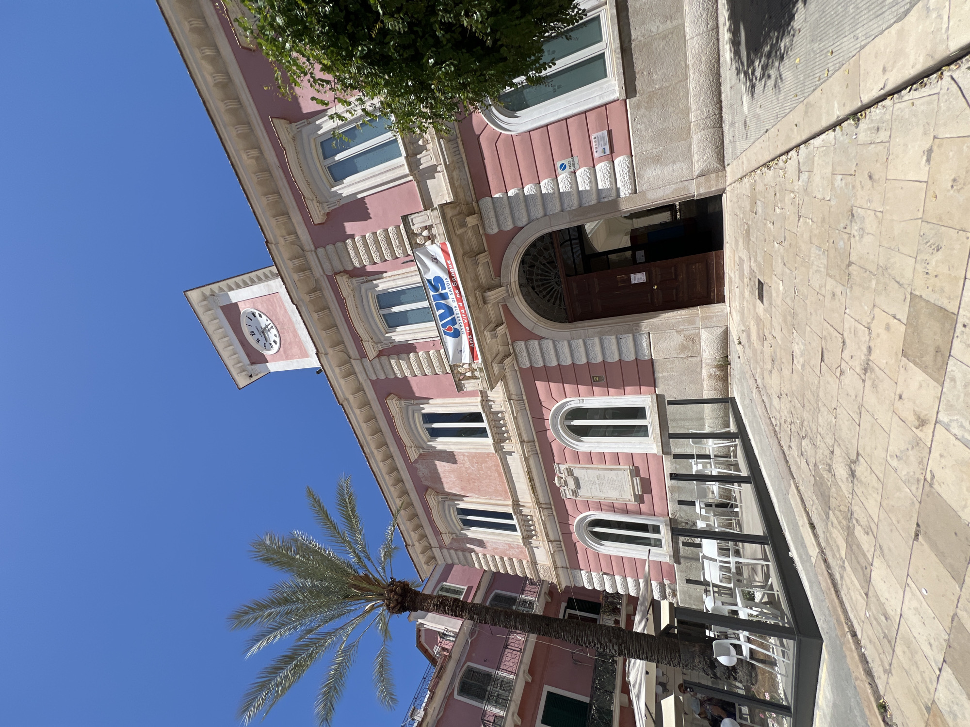 The image depicts a street scene in Margherita di Savoia, Italy. The primary subject is a pink building with white trim and ornate architectural details, including arched windows and decorative stonework. A clock tower, also part of the building, is visible on the left, showing Roman numerals on its face. A palm tree stands in the foreground, its fronds reaching upwards towards a clear blue sky. The street is paved with large stone tiles.
In front of the building, there is an outdoor seating area with white tables and chairs, suggesting a cafe or restaurant. A banner hangs from the building, displaying text in Italian, including "AVIS" and other phrases that are partially obscured. Signs are also visible on the building's facade, one with a blue and white symbol.
The time of day appears to be daytime, with bright sunlight casting shadows. The weather seems clear and sunny. The overall mood is serene and characteristic of a Mediterranean town. The image captures a slice of everyday life in an Italian town, blending architecture, nature, and public spaces.