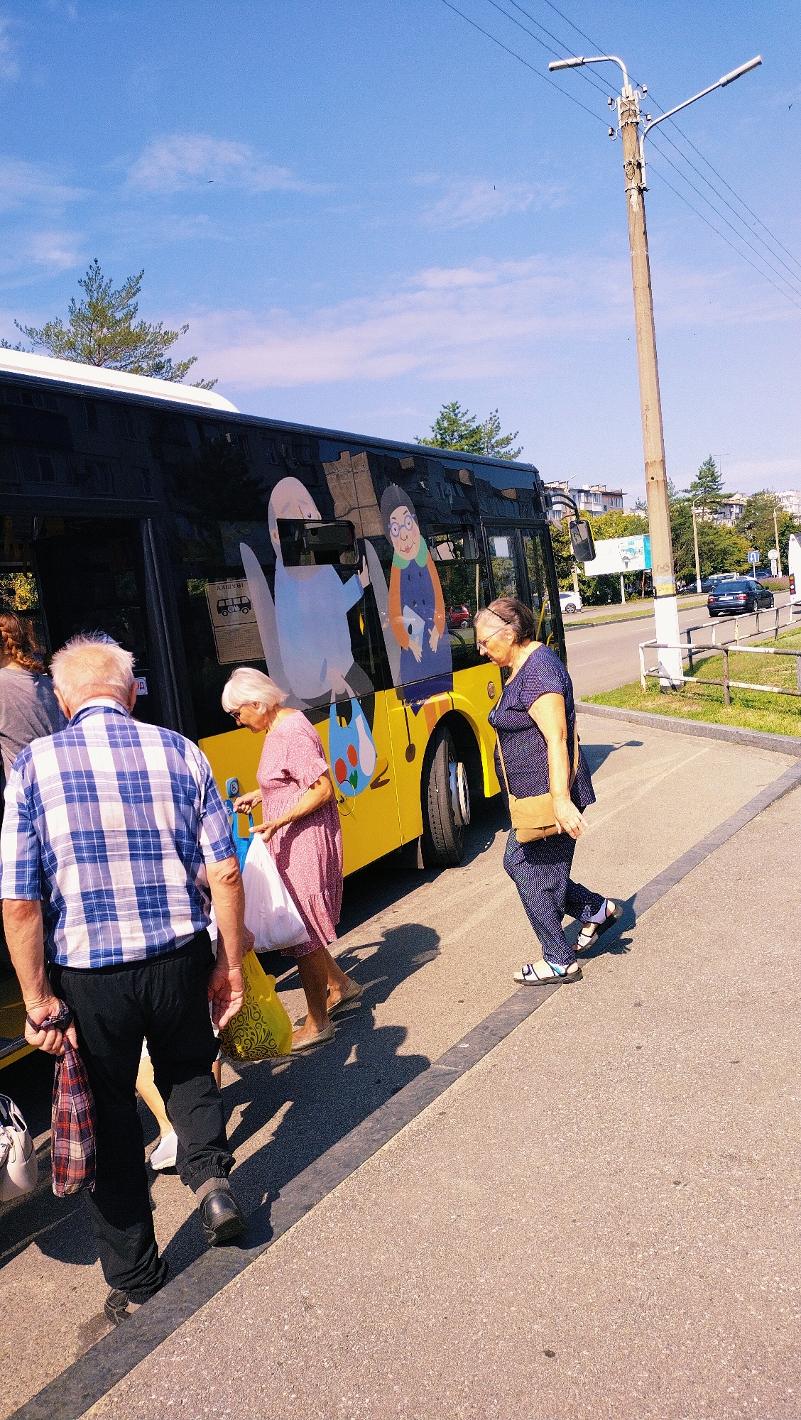 In Horishni Plavni, Ukraine, a bright yellow bus with cartoonish illustrations on its side is parked at a bus stop on a sunny day. Several people are gathered around the bus, some appearing to be boarding or disembarking. In the foreground, a man in a blue and white plaid shirt and black pants is walking away from the bus, carrying a patterned bag. To his left, a woman in a pink dress with white trim is holding shopping bags, one of which is white and the other yellow. Further to the right, a woman in a dark blue patterned outfit with a tan purse slung across her body is walking away from the bus. The bus features a large cartoon depicting an elderly woman with glasses and a blue top, as well as another cartoon of an elderly person. A utility pole with multiple streetlights and wires stands prominently near the bus stop. The environment appears to be a street with a sidewalk and a road in the background, where a dark sedan is visible. Trees and buildings are present in the distance.