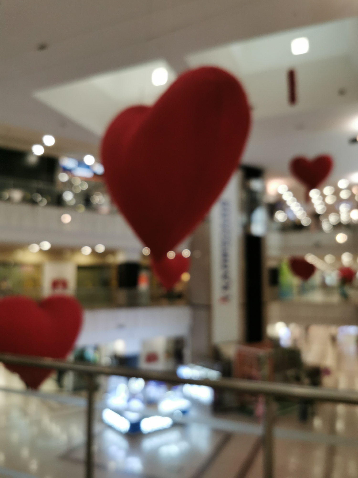The photograph shows a blurred indoor scene, likely a shopping mall in Moscow, Russia based on general architectural styles of malls in the city. Several large, red, heart-shaped decorations are suspended in the air. A metal railing is visible in the foreground. In the background, multiple levels of the mall can be seen, with shops and bright lighting. People are vaguely discernible in the blurry background, but their actions and interactions are not clear.
