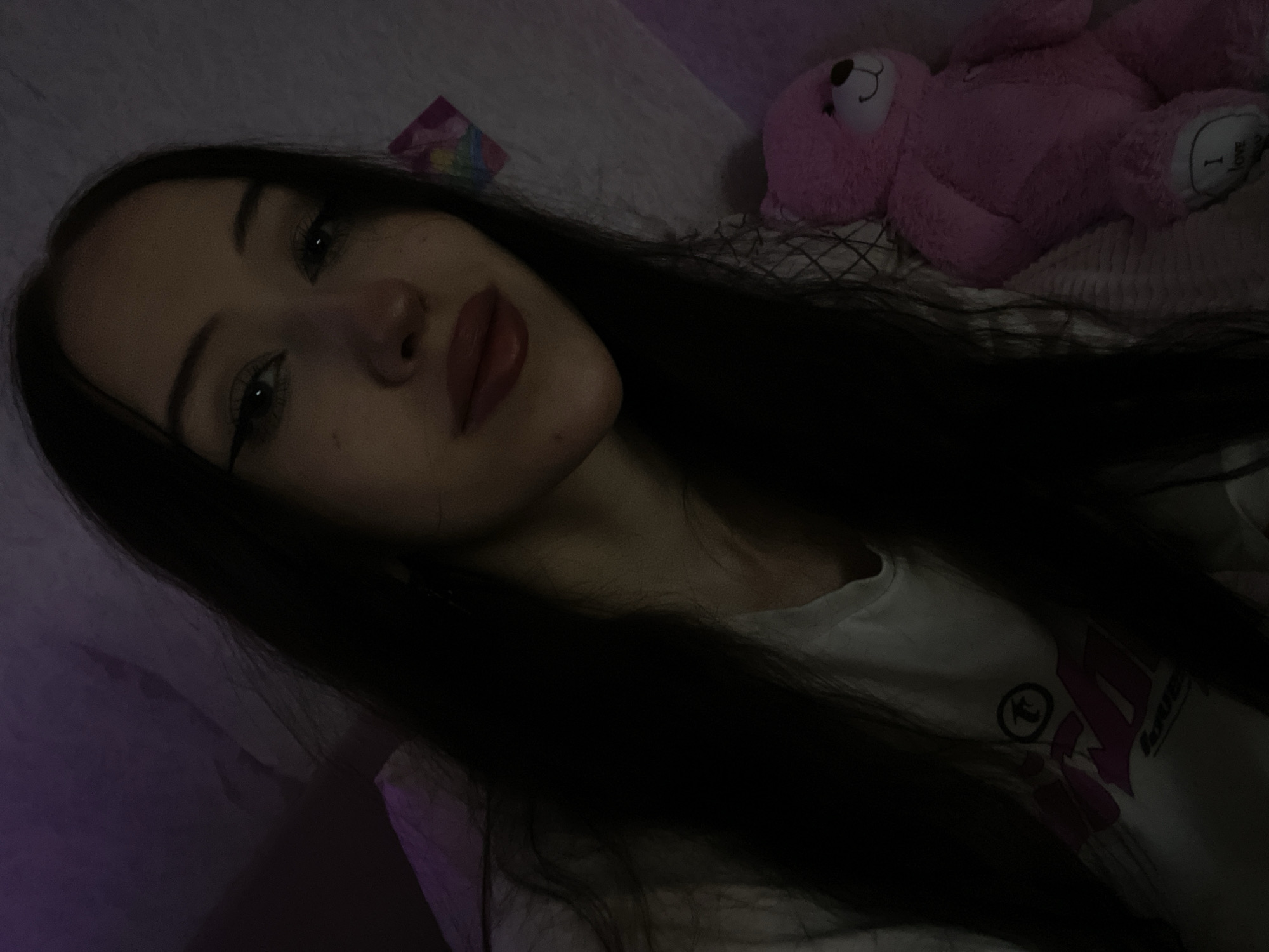 The image is a selfie taken indoors, likely in a bedroom, given the soft lighting and presence of a stuffed animal. A young woman with long, dark hair and winged eyeliner is the primary subject. She is wearing a white t-shirt with graphics on it. To her right, a pink stuffed teddy bear is visible, propped up against a surface. The background appears to be a wall with some decoration, possibly stickers, and a patterned texture that could be bedding or a wall covering. The lighting is dim and has a purple hue, suggesting it might be from a colored light source or low-light conditions. There is no indication of the location being in Votkinsk, Russia.