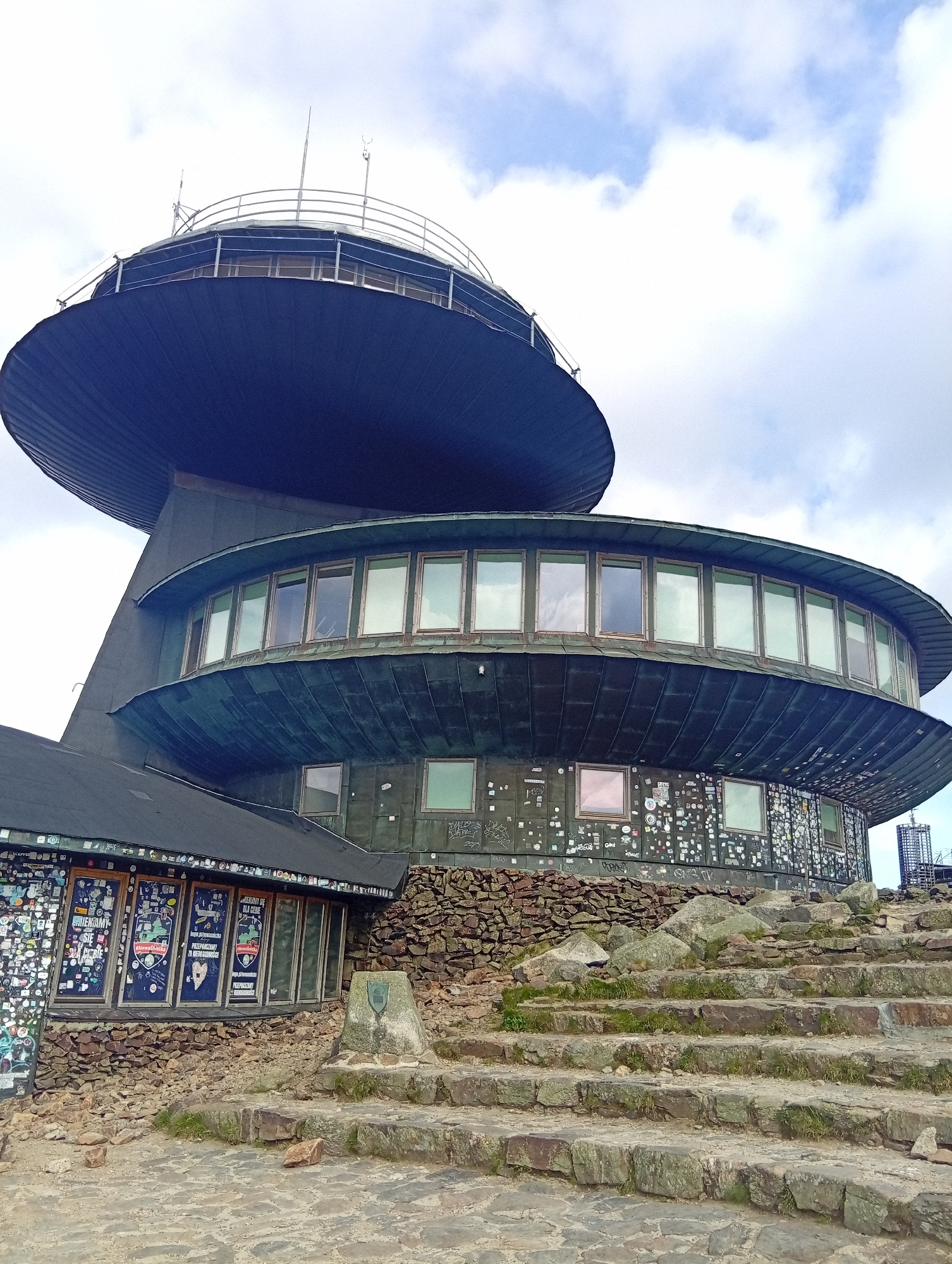 The image depicts the Śnieżka mountain observatory, a distinctive building situated atop the mountain of the same name, which straddles the border between the Czech Republic and Poland. The observatory is characterized by its two circular, stacked structures. The upper structure is a large, dark, disc-like element with a railing around its perimeter and what appear to be antennas or meteorological instruments on top. Below this is a wider, also circular section with numerous large windows, reflecting the sky. The lower part of the building, which is more angular, has a dark roof covered in a multitude of stickers and posters, and its walls are also adorned with many stickers and some graffiti. This section is built into a rocky hillside, with a stone-clad base. In the foreground, a set of uneven stone steps leads up towards the building. The ground is paved with irregular stones and scattered rocks. The sky is partly cloudy with patches of blue and white. There are no people visible in the image. The overall impression is of a unique architectural landmark in a high-altitude, rugged environment.
