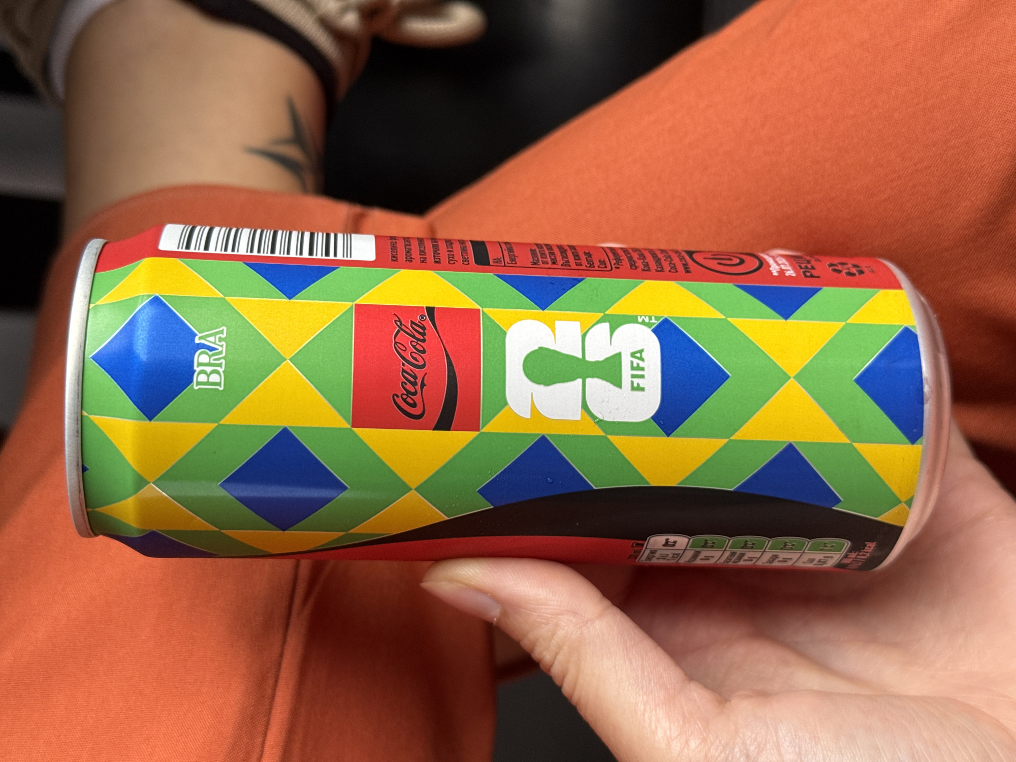 A left hand holds a Coca-Cola beverage can vertically. The can features a commemorative design with a repeating pattern of green, yellow, and blue diamond shapes, evoking the Brazilian flag. White text "BRA" is positioned near the top, above the classic red Coca-Cola logo. Below the logo, a white silhouette of a football player holding a ball (representing the FIFA World Cup trophy) is centrally placed, with the word "FIFA" in white text beneath it.
On the right side of the can, visible white Cyrillic text is present, indicating a Slavic language, adjacent to a barcode. Partially visible on the left side of the can is nutritional information in white text on a black background, showing values like "0g" and "0 kcal / 13 kj" per 100 ml. Small droplets of condensation are visible on the can's surface, suggesting it is cold.
The hand holding the can is light-skinned, with the thumb pressed against the left side. The immediate background underneath the can and hand is a textured, orange-colored fabric, likely clothing. In the blurred background to the right of the can, a portion of a human leg or hip is visible, featuring a dark, abstract tattoo. Further blurred elements, including a striped pattern, are discernible in the upper right. The overall environment is indistinct and appears to be indoors with soft lighting.
The image itself does not contain any visual information to confirm a specific location within the city of Pleven, Bulgaria.