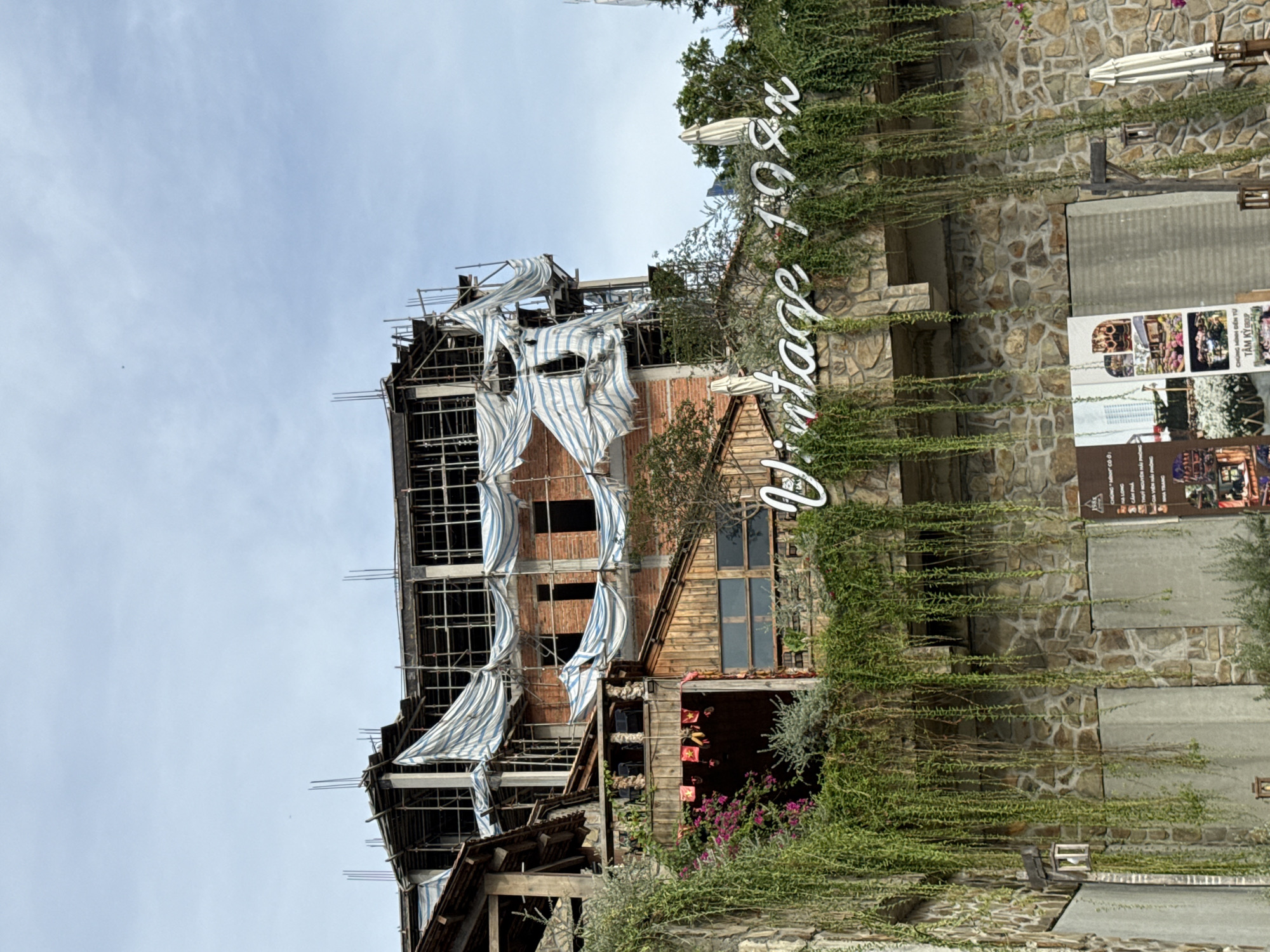 This is an eye-level, medium shot of a building under construction in Nha Trang, Vietnam, with a sign that reads "Vintage 198x" prominently displayed. The building is a multi-story structure with exposed brickwork and scaffolding covered in blue and white tarps. Lush green vines cascade down the stone facade of the lower section of the building, partially obscuring windows. A stone wall forms the base of the structure. To the left, a balcony area with wooden railings and chairs is visible. Red and yellow banners with Vietnamese stars hang above an entrance. In the lower right corner, an umbrella and more greenery are present. The sky is cloudy and light blue.