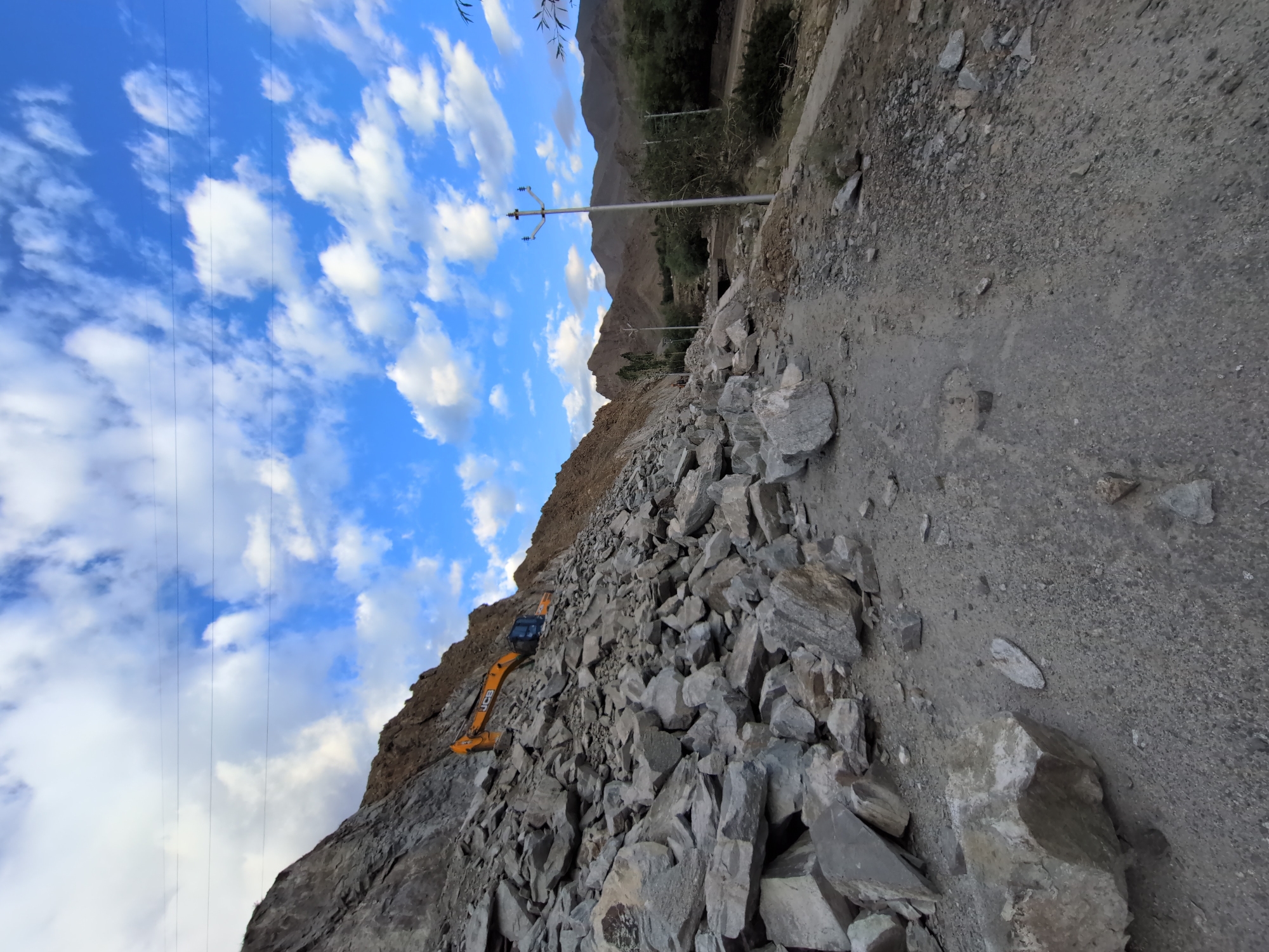 The image captures a construction site near the city of Leh, India. A large excavator, predominantly yellow and black, is positioned on a steep, rocky slope. The slope is covered with numerous grey rocks and debris, suggesting ongoing excavation or landslide clearing. The sky above is a vibrant blue, dotted with fluffy white clouds. In the mid-ground, a power line pole with multiple insulators stands prominently, with more poles and lines visible in the distance. Further back, mountainous terrain forms the backdrop, with some trees and vegetation visible in the lower valley area. A dirt or gravel road runs along the foreground and into the mid-ground, with scattered rocks and debris on its surface. The scene appears to be in a rugged, mountainous environment, characteristic of the region around Leh. There are no people visible in the image.