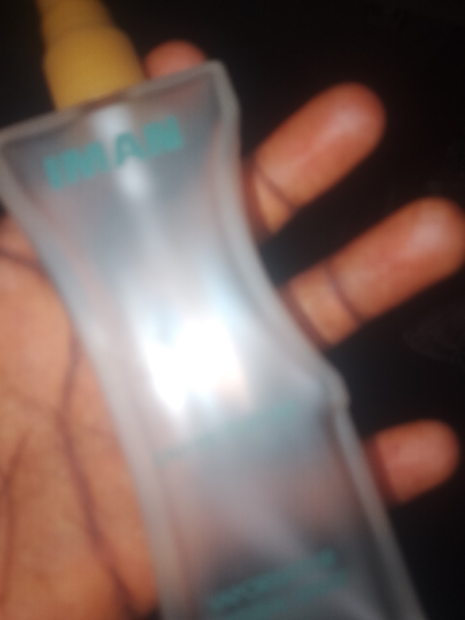 A slightly blurry, close-up, vertical shot shows a person's hand holding a frosted, transparent plastic bottle with a yellow cap. The bottle has teal-colored text on it, which appears to read "IMAN." The background is dark and out of focus, with some reddish-orange highlights likely from lighting. The image does not contain any information that can be used to determine its location in Jauro Yinu, Nigeria, or any other specific geographical location.
