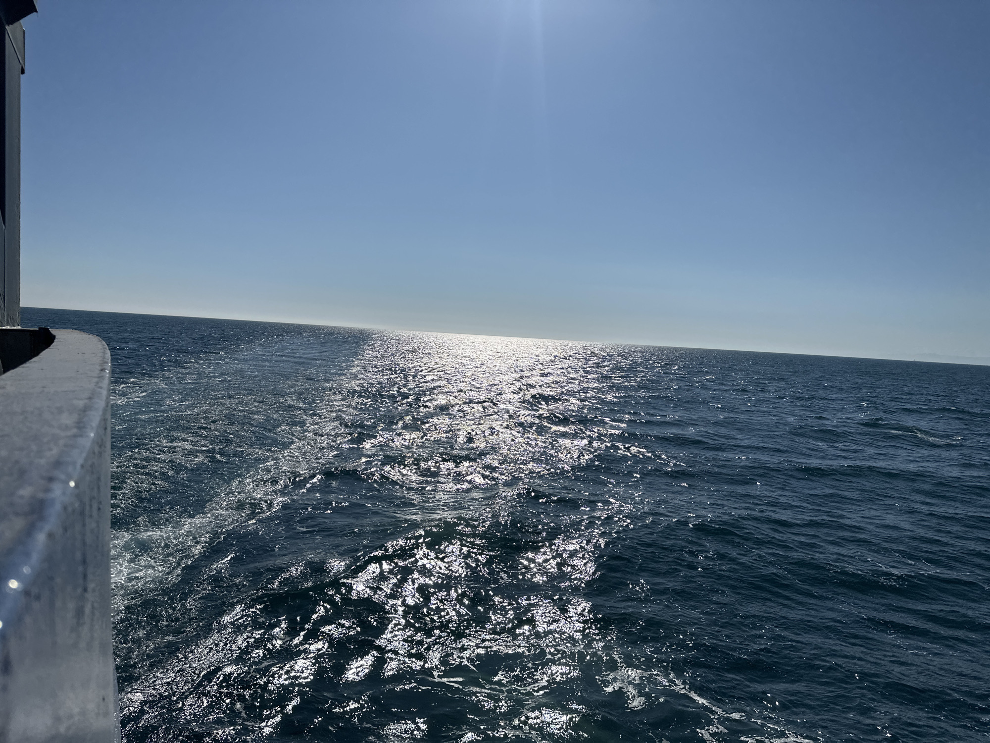 The image shows a seascape under a bright, clear sky, with the sun's glare reflecting off the water. The dark blue water is choppy, exhibiting a wake of froth and bubbles closest to the viewer's vantage point, presumably from a moving vessel. A small section of the vessel's railing or structure is visible in the lower left corner. The horizon is distinct, with a landmass faintly visible on the right. The location is at sea, within the city of Unknown, Unknown.