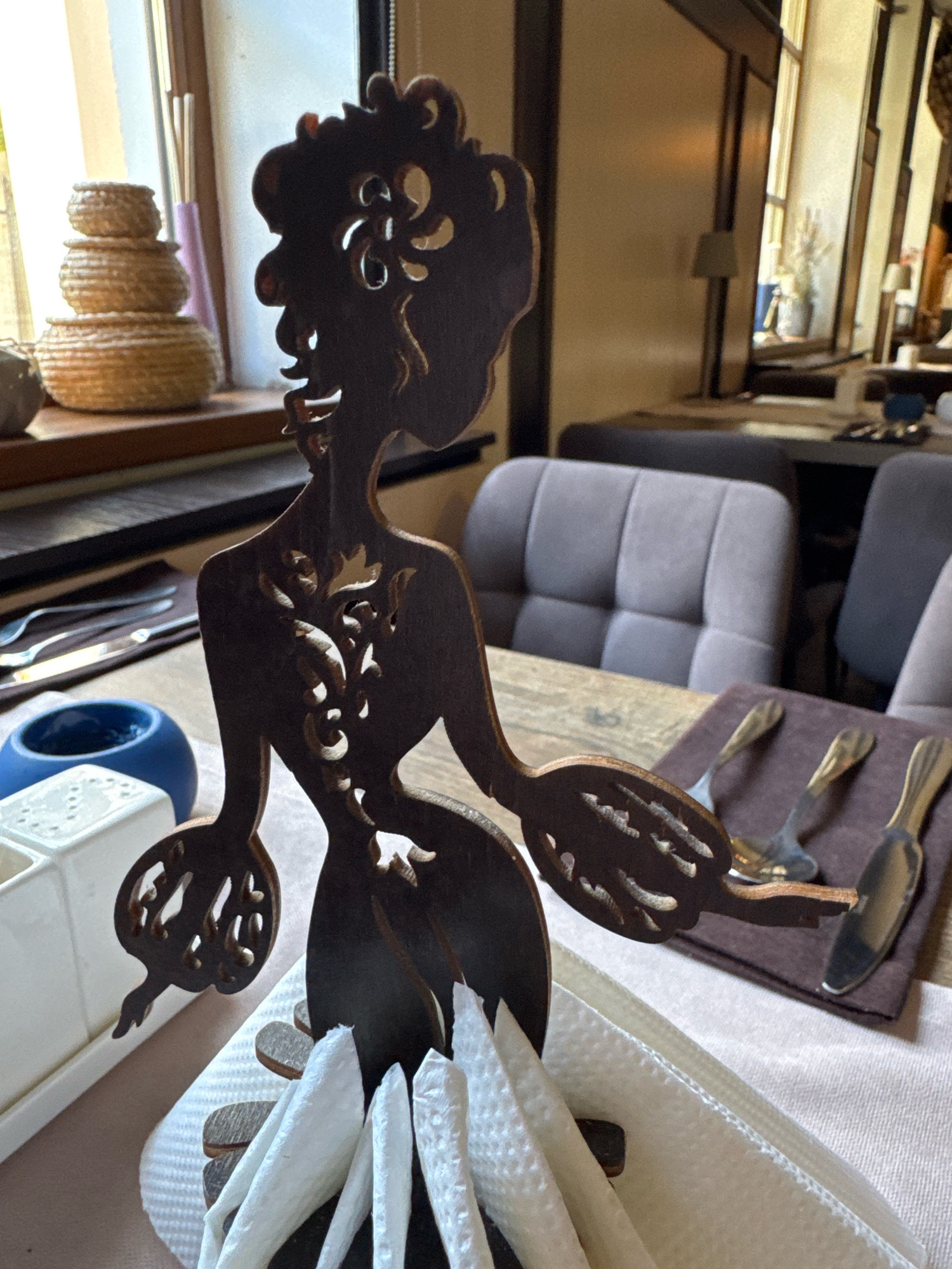 The image depicts a close-up of a wooden silhouette of a woman, likely a decorative piece, standing on a table. The silhouette has intricate cutouts in its torso and arms. It is positioned in front of a white napkin holder filled with white napkins. To the left of the silhouette, a small blue ceramic dish and a white rectangular container are visible. In the background, a window with light streaming through can be seen, with stacked woven baskets on the windowsill. Further in the background, a restaurant or cafe setting is apparent with tables, upholstered chairs, and dimly lit lamps. The table is set with cutlery on a brown cloth. The overall environment suggests an indoor dining establishment. There are no people or animals visible in the image. The location is not identifiable from the image itself.