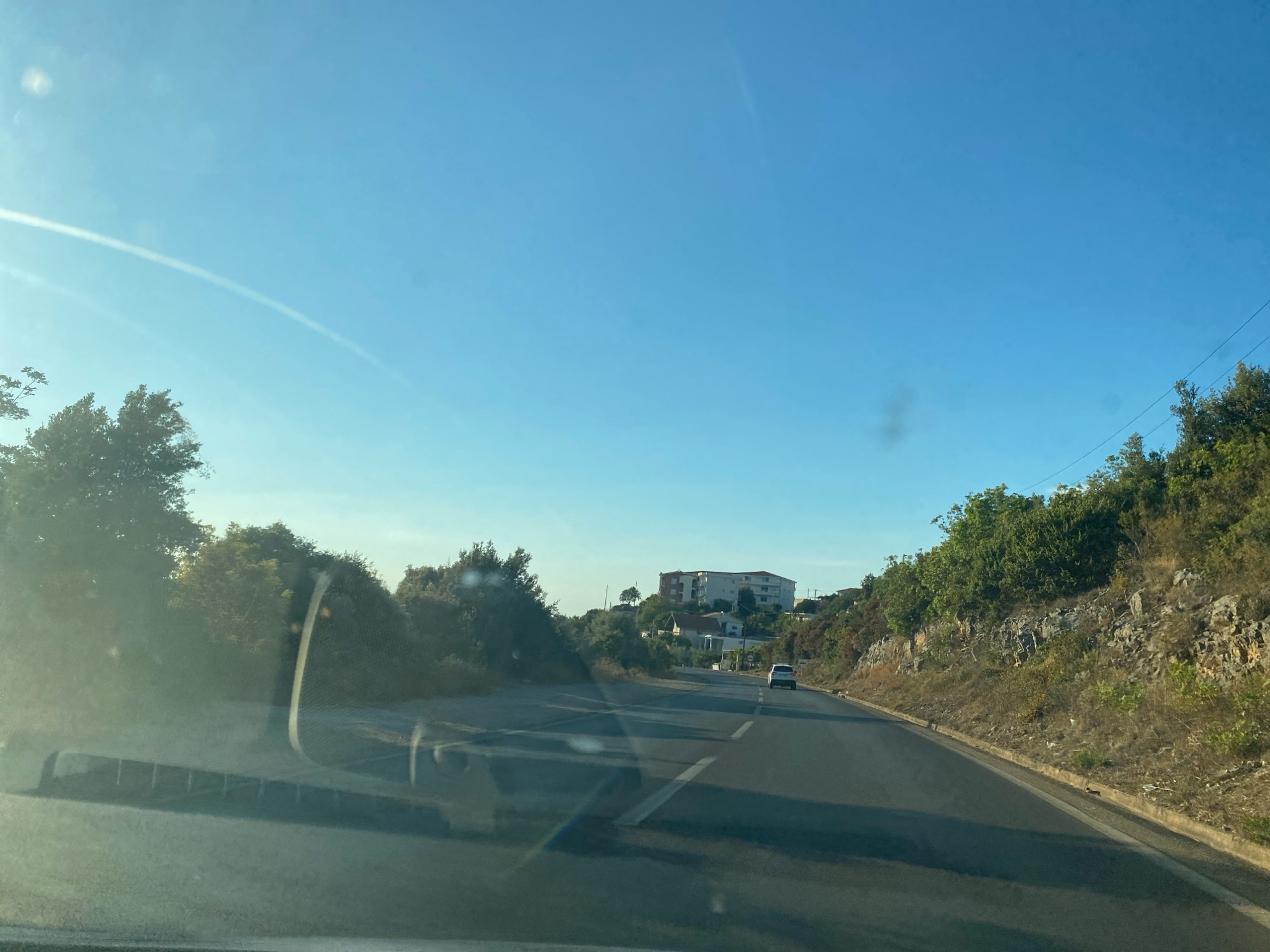 This is a first-person perspective shot from inside a vehicle traveling on a paved road in Kunje, Montenegro. The road curves slightly to the right as it ascends a gentle hill. On the left side of the road, dense green foliage lines the shoulder, with scattered trees. The right side of the road features a rocky, uneven embankment covered with dry vegetation and scattered trees. In the distance, atop the hill, several buildings are visible, including a large, multi-story structure and smaller houses. A single white car is traveling in the same direction as the viewer, ahead on the road. The sky is clear blue with a faint contrail extending diagonally from the upper left to the center of the frame. The sunlight creates a glare on the windshield, with visible streaks and smudges.