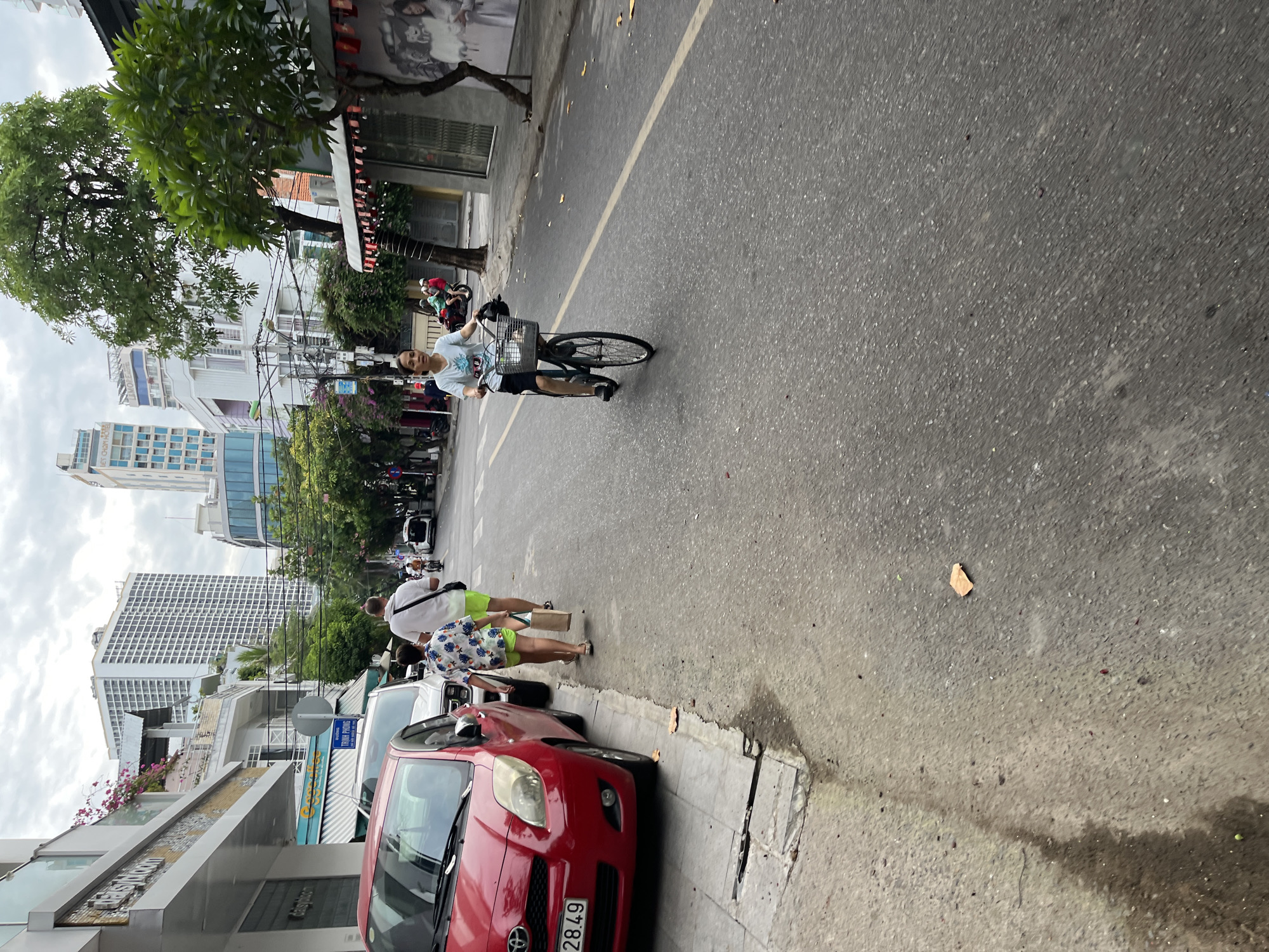 This image depicts a street scene in Nha Trang, Vietnam. On the left side of the road, a red Toyota Yaris car is parked along a concrete sidewalk. Further down the sidewalk, two people are walking away from the camera: a man in a white t-shirt and green shorts, and a child in a patterned shirt and green shorts, carrying a brown paper bag.
The street itself is paved with asphalt and has a yellow dividing line. A woman is riding a bicycle towards the camera on the right side of the road. She is wearing a light-colored t-shirt and dark shorts, and a basket is attached to the front of her bicycle. In the background, several multi-story buildings, including a tall white building with many windows, are visible, suggesting an urban environment. Green trees line the street, and there are some overhead power lines. The sky is overcast. The scene appears to be an ordinary day in the city.