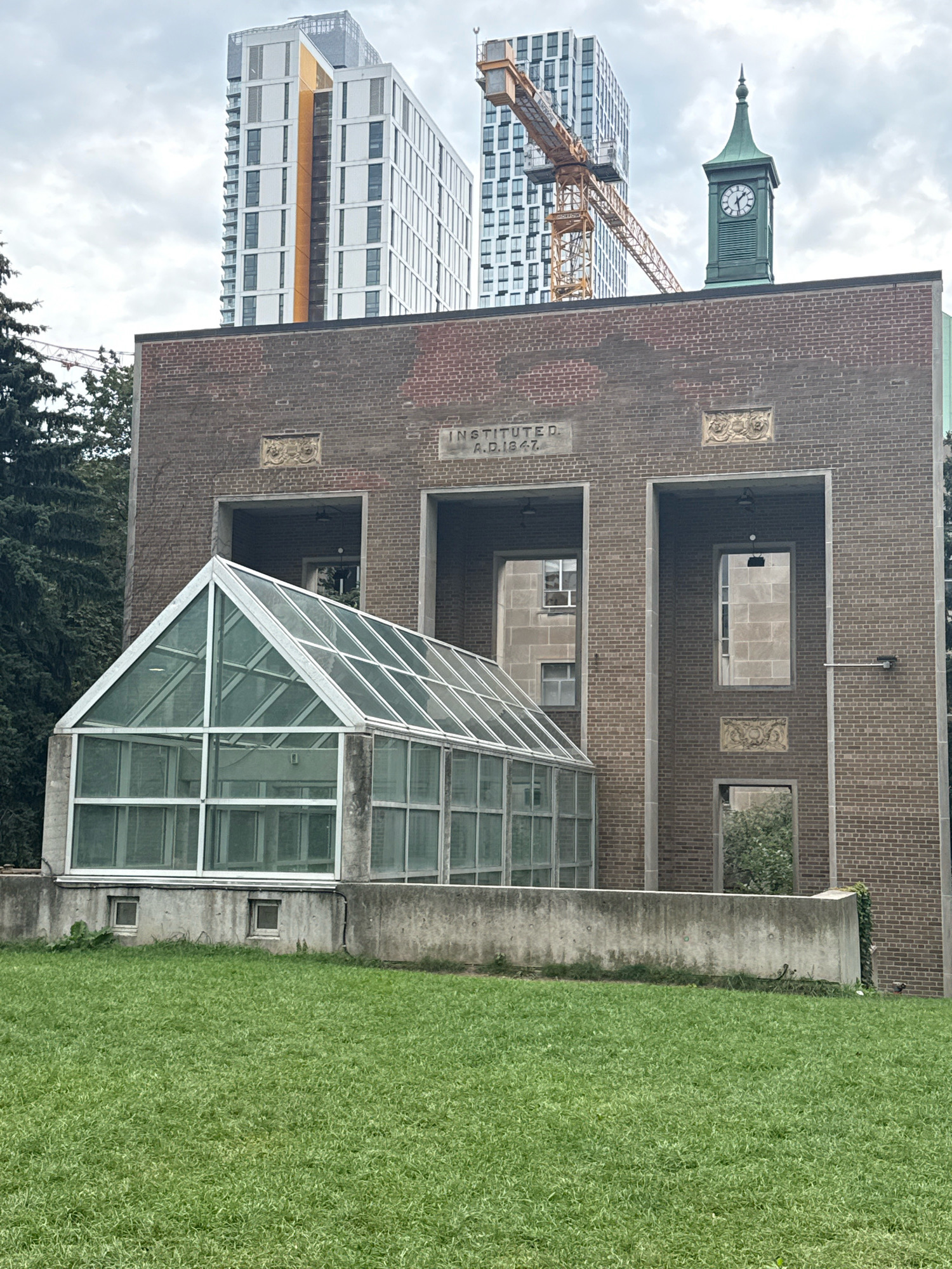 The image shows the exterior of the Royal Ontario Museum's Institute for Quantitative Analysis in Toronto, Canada. The building features a traditional brick facade with large, recessed archways and decorative stone carvings. Attached to the front of the building is a modern glass greenhouse with a white metal frame. In the background, tall, contemporary skyscrapers and a construction crane rise above the older structure. A green clock tower with a pointed spire is visible atop the brick building. The foreground consists of a lush green lawn. The sky is overcast.