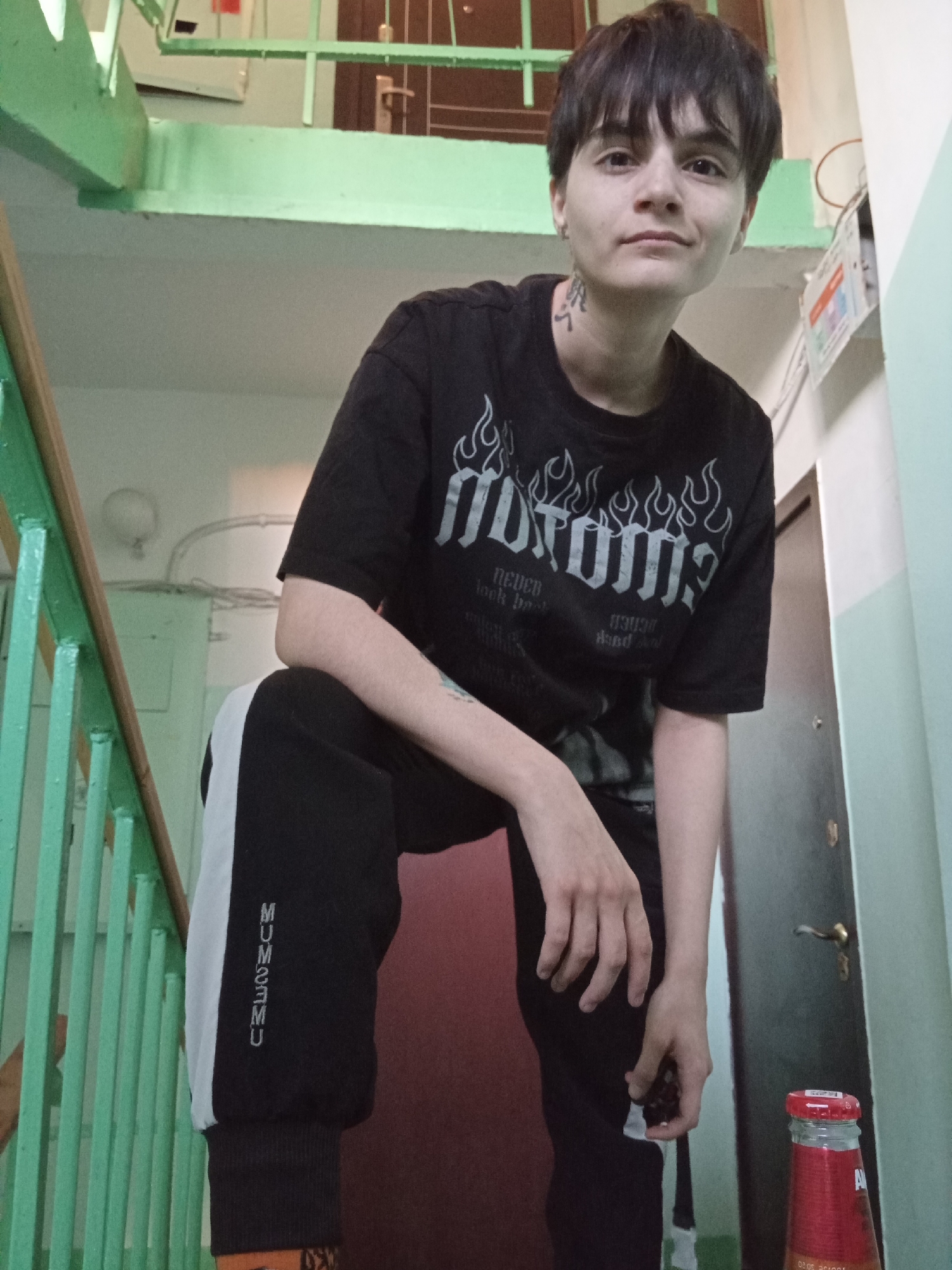 A person with short, dark hair is posing in a stairwell. The person is wearing a black t-shirt with a white graphic design and black sweatpants with a white stripe down the side, upon which is the reversed word "MUMUMUMU". The person is standing on a lower step with one knee bent and the other leg extended, while holding onto a railing of green-painted metal. A bottle of soda with Russian writing is visible in the lower right corner of the frame. The stairwell is located in Naberezhnye Chelny, Russia, as inferred from the Cyrillic text on the soda bottle. The environment includes painted walls, a door, and utilitarian features of a public building like lighting fixtures and posters.