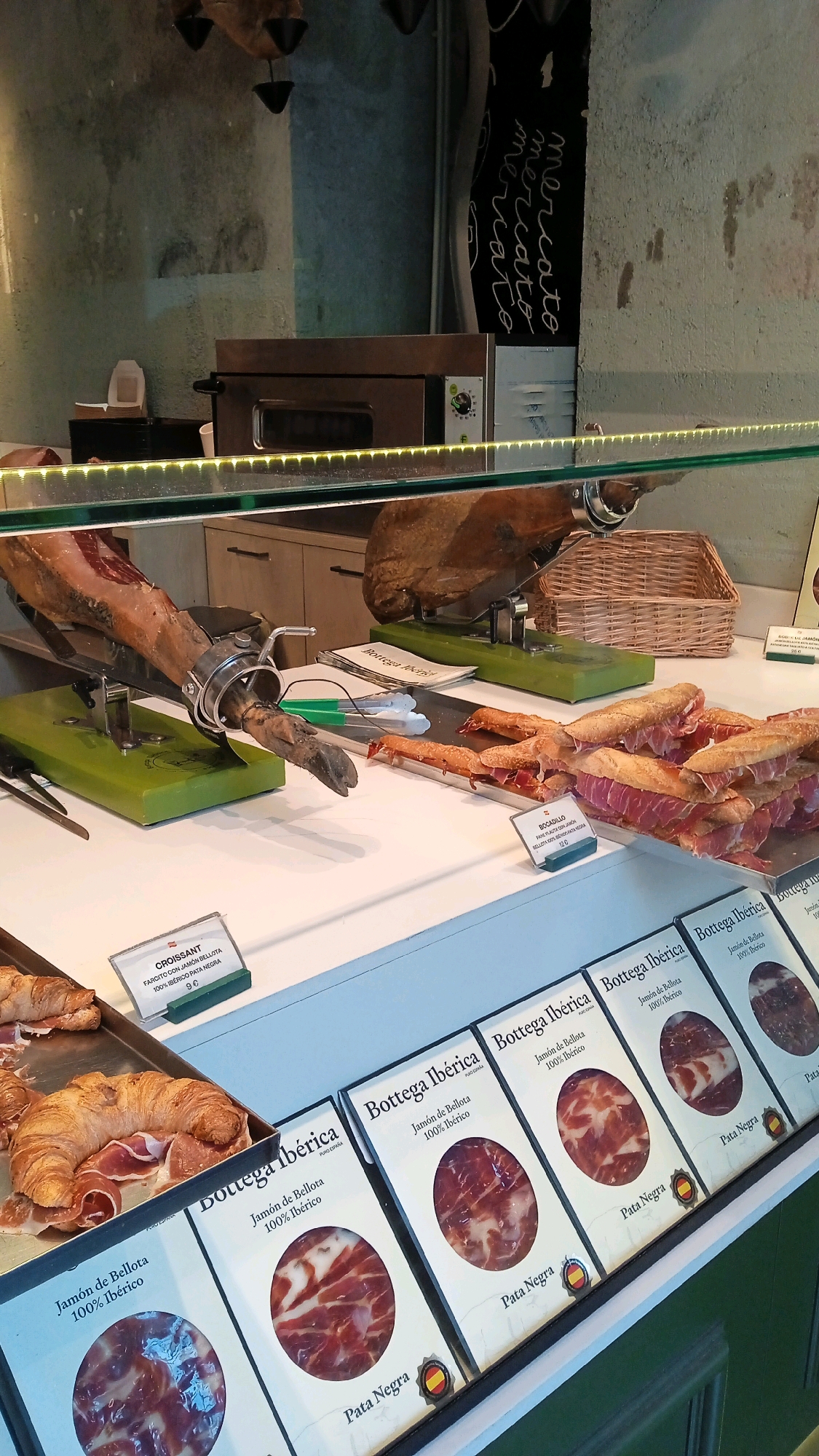 The image shows a display case filled with various Spanish hams and pastries, likely in a deli or market in Milan, Italy, given the "Mercato" inscription on the wall. In the foreground, a large leg of cured ham is presented on a green cutting stand, with a few slices already cut and placed on a metal tray to its right, alongside what appear to be bocadillos (sandwiches). Further to the right, a tray of croissants with ham is visible. The background reveals a commercial oven and a wicker basket. The display case is illuminated by strip lighting, highlighting the food items. The overall setting suggests a food establishment focused on high-quality Iberian products.