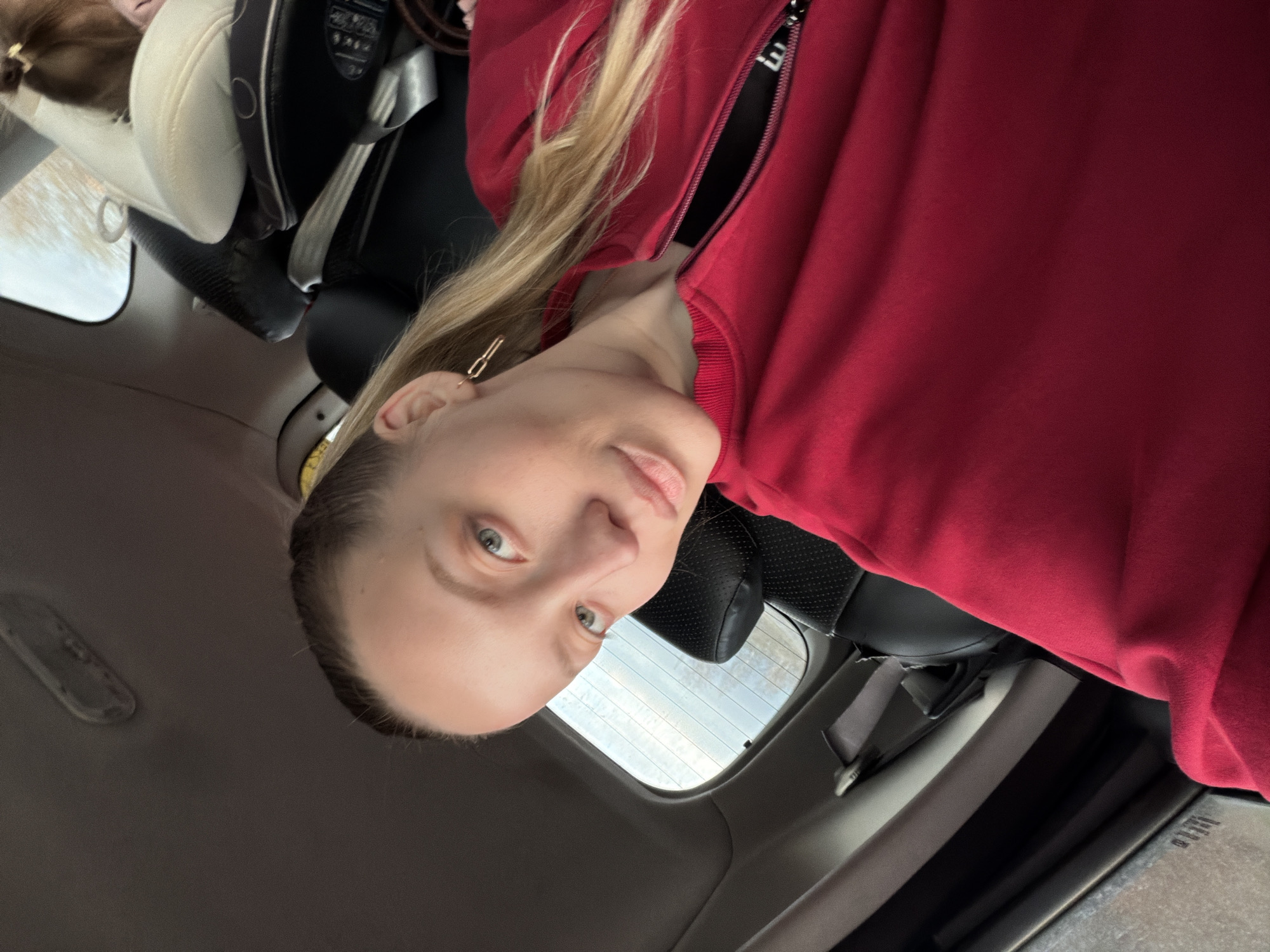 A woman with blond hair and blue eyes is in the back seat of a car. She wears a red zip-up sweater and a gold hair clip. A car seat is beside her. Outside the car window, the sky is visible. The location is assumed to be Razdol'e, Russia.