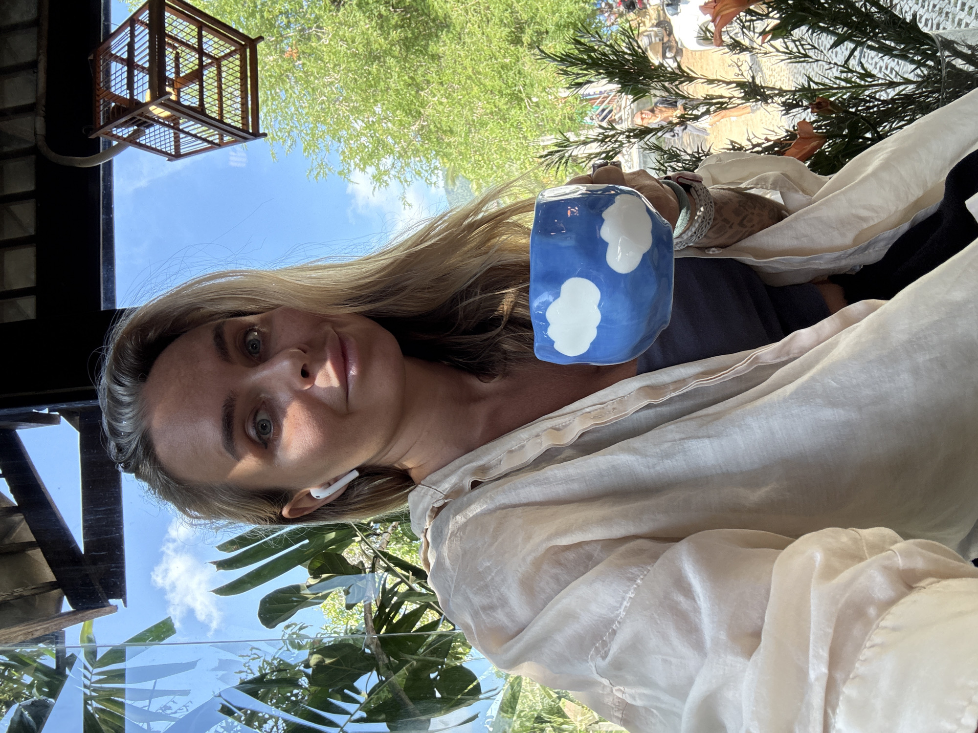 A woman with blonde hair and blue eyes is taking a selfie. She is wearing a beige, open button-up shirt over a dark top, and white wireless earbuds. She is holding a blue ceramic mug with white cloud designs. The woman is seated indoors, with a large window behind her that overlooks a lush green outdoor environment with trees and a bright blue sky. Through the window, a glimpse of a wooden birdcage hanging from the ceiling is visible, as well as distant mountains and some people. She appears to be in a cafe or restaurant setting. The image was taken at The Lazy Bar cafe, located in the Bophut district of Koh Samui, Thailand.
