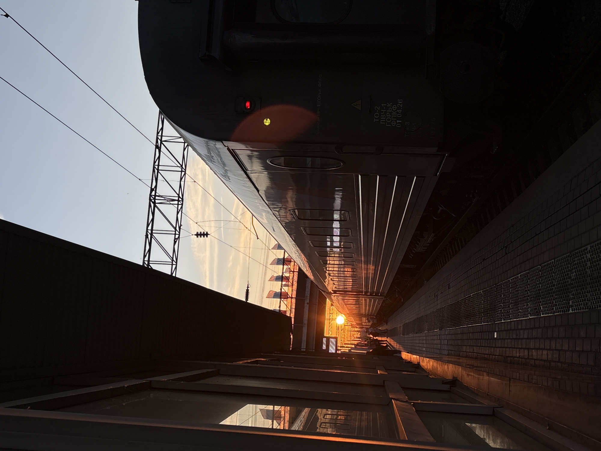 The image captures a sunset at a train station in Volgograd, Russia. A train is parked on the tracks, its dark carriages contrasting with the warm hues of the setting sun. The sun is a bright orb low on the horizon, casting long shadows and illuminating the platform with golden light. On the left side of the image, a covered platform area with windows reflects the scene. A few people are visible on the platform, some walking, others appearing to wait. In the background, several Russian flags are displayed on poles, hinting at the location. Overhead, power lines and a section of overhead track infrastructure are visible against the pale sky. The platform is paved with bricks, and the train's wheels and lower structure are visible near the tracks. The overall atmosphere is serene, with the end of the day casting a tranquil glow over the urban setting.