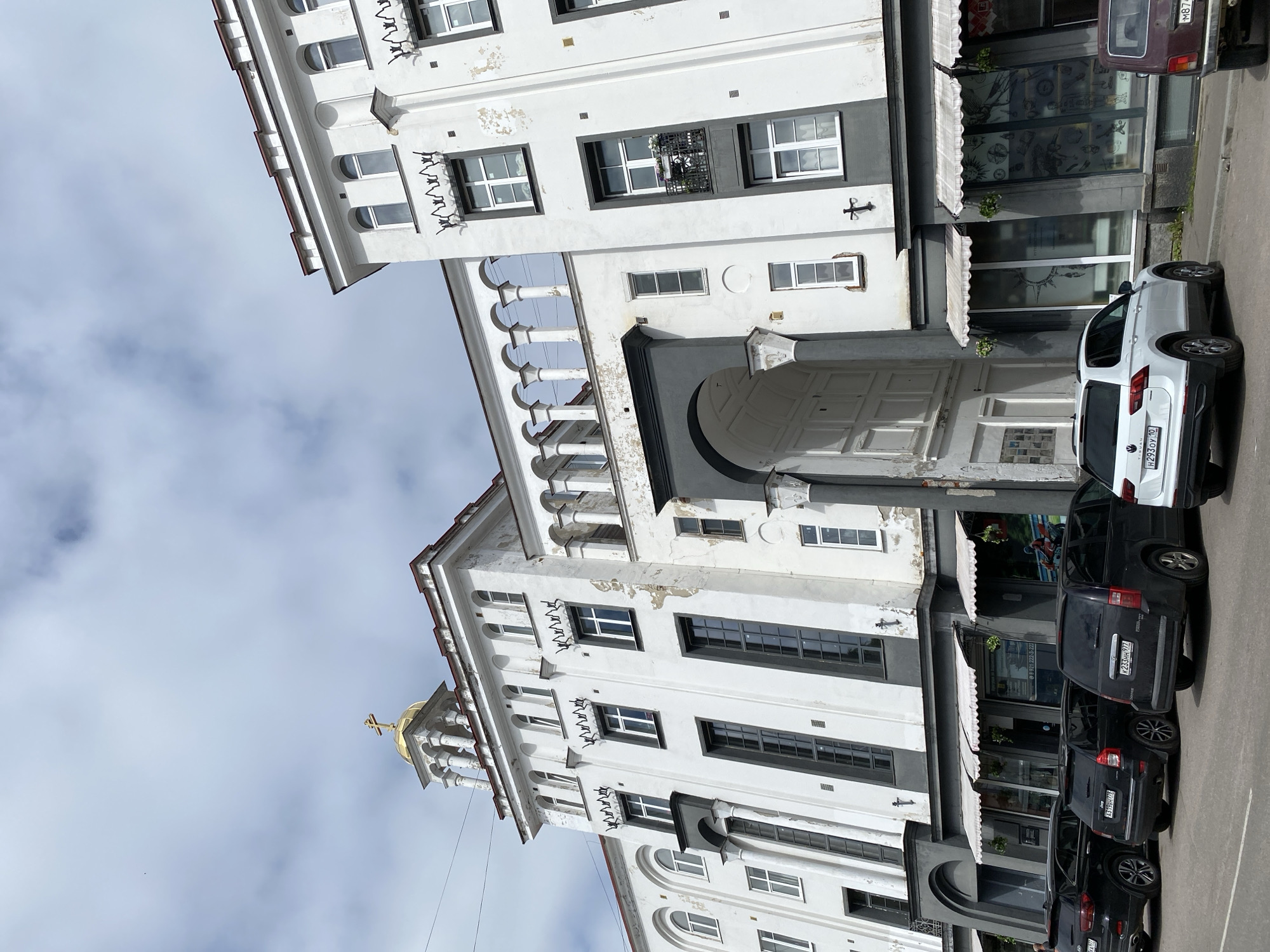 The image shows a white building with grey accents in Sortavala, Russia, featuring a distinctive golden dome with a cross at its peak. The building has an arched entryway and a colonnade on the upper levels. Several black cars are parked in front of the building, along with one white SUV. The sky is overcast with clouds. The building appears to be commercial or mixed-use, with shopfronts visible on the ground floor. The facade of the building exhibits some signs of wear and tear, with peeling paint in places.