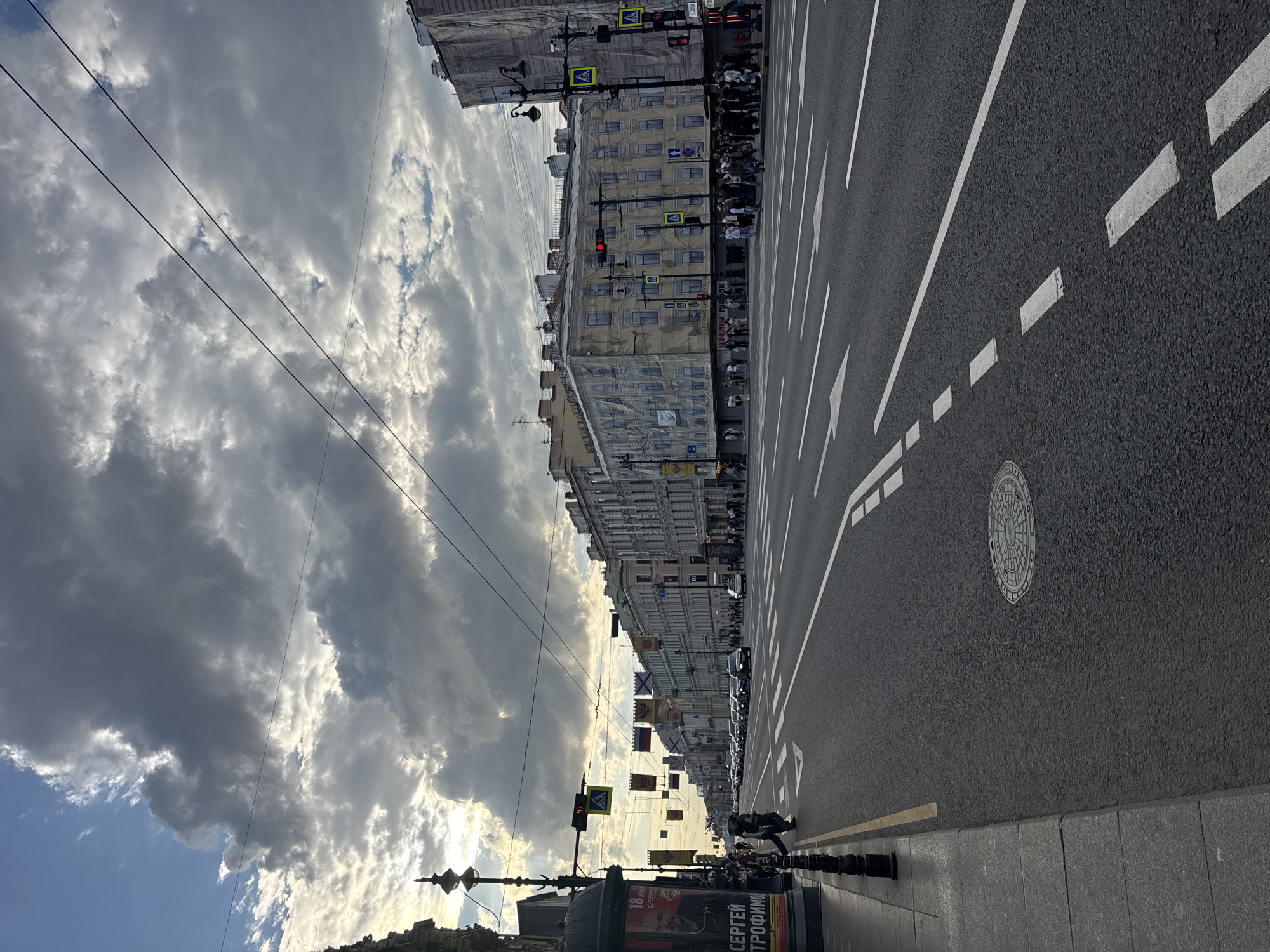 This street-level photograph captures a dynamic urban scene in Saint Petersburg, Russia, likely during the late afternoon or early evening given the lighting. The sky dominates the upper portion of the image, filled with dramatic, heavy cumulus clouds that are brightly illuminated by the sun breaking through, creating a contrast with darker cloud masses. Patches of blue sky are visible towards the lower left. Overhead, multiple power lines or trolley lines traverse the sky.
The main subject is a wide asphalt road, clearly marked with white lane lines, dashed lines, and directional arrows. A circular manhole cover is visible on the road surface. To the right, a sidewalk runs parallel to the street, featuring a black decorative street lamp and dark bollards. A large cylindrical advertising stand stands prominently on the sidewalk in the foreground.
Along the street, multi-story, classical-style buildings line the left side. Several of these historical structures are covered with yellow construction netting or printed facades, suggesting ongoing renovation or repair. Pedestrian crossing signs (yellow diamond with black person symbol) are visible on poles, as well as traffic lights, with one showing a red signal and another displaying a "41" countdown timer.
Numerous people are visible on the sidewalks, some walking, others potentially waiting at crosswalks. Cars are parked or moving in the distance along the street, and a motorcycle or scooter is parked near the sidewalk in the mid-ground. Decorative banners or flags, some resembling naval ensigns, are strung across the street between buildings, adding to the urban ambiance.
Visible text on the advertising stand reads "18 МАЯ" (May 18th) and "СЕРГЕЙ ТРОФИМОВ," likely promoting an event or concert. Another building facade shows partially obscured text, including "ЦВЕТЫ..." (Flowers...). The scene depicts typical city life under a striking sky, with elements of everyday transport and urban development.