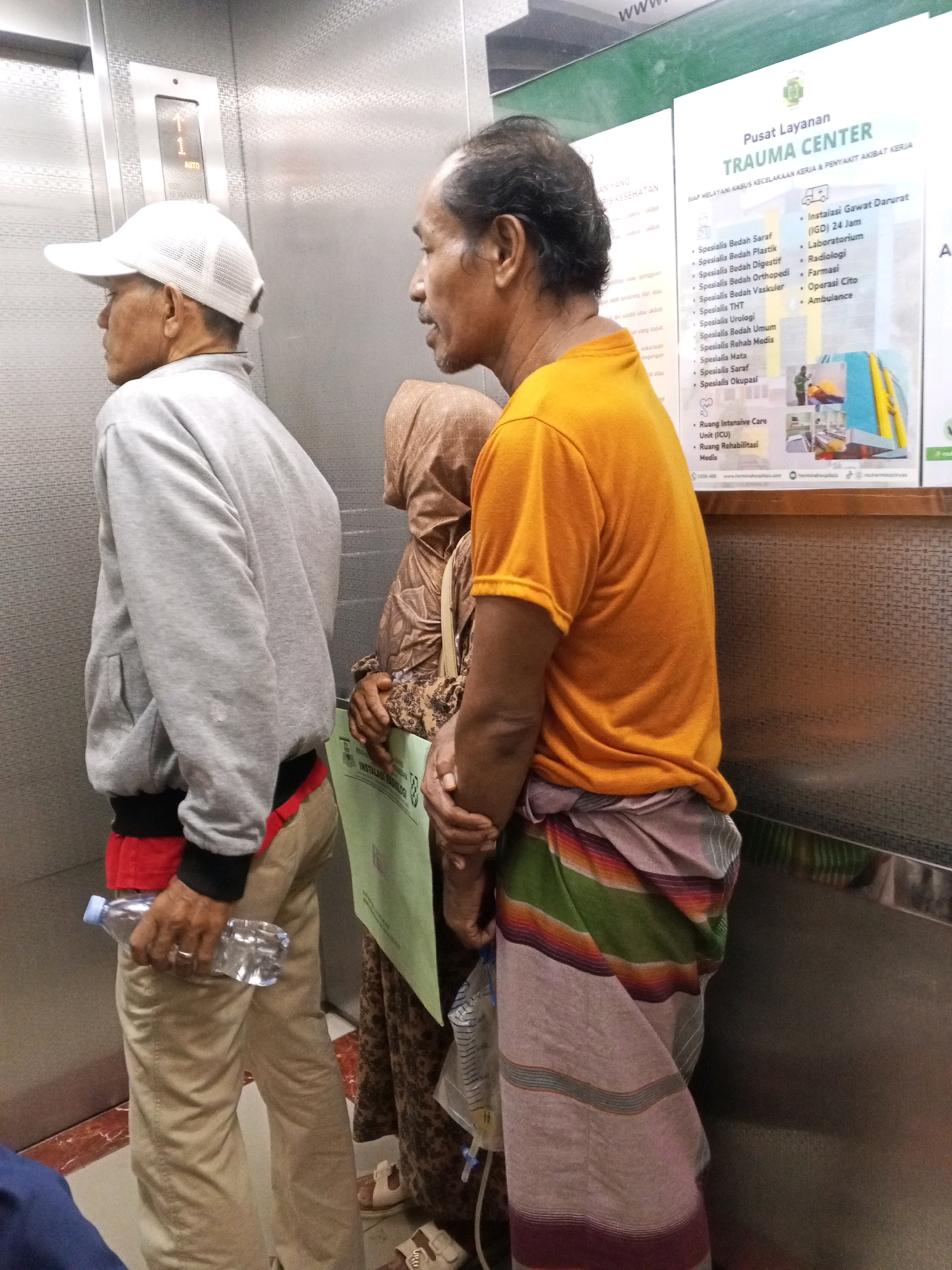 The image shows three people in an elevator with metal walls. A man in a white baseball cap and gray sweatshirt is on the left, holding a clear plastic water bottle. In the center, a person in a brown hijab and patterned dress is visible. On the right, a man in an orange t-shirt and a sarong with horizontal stripes in green, red, and purple is looking to the left. Behind the people, there is a poster in Indonesian for "Pusat Layanan TRAUMA CENTER," which appears to be a trauma center service. The poster lists various medical specialties and services, including "Instalasi Gawat Darurat (IGD) 24 Jam" (Emergency Room 24 Hours), "Laboratorium" (Laboratory), and "Ambulance." The location is identified as Ciruas, Indonesia.