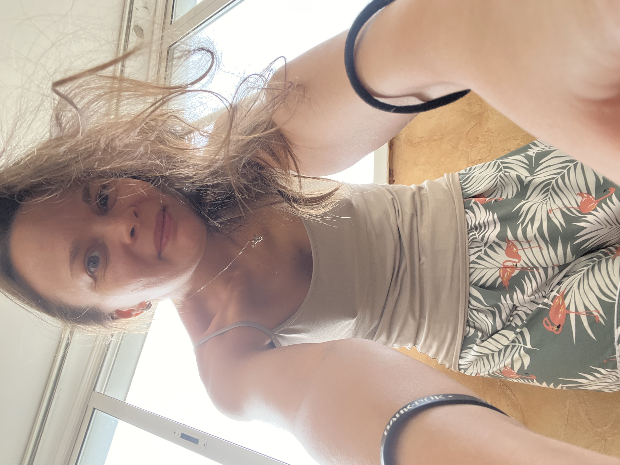 A woman with long, light brown hair is taking a selfie, looking down at the camera. She is wearing a light beige tank top and patterned shorts with palm leaves and flamingos. A black bracelet is visible on her left wrist. Her hair is blowing around her face, suggesting an outdoor setting or a strong breeze. The background shows a window with bright daylight streaming in, indicating it is daytime. The overall scene appears casual and relaxed. The location is Tolyatti, Russia. There is no visible text.