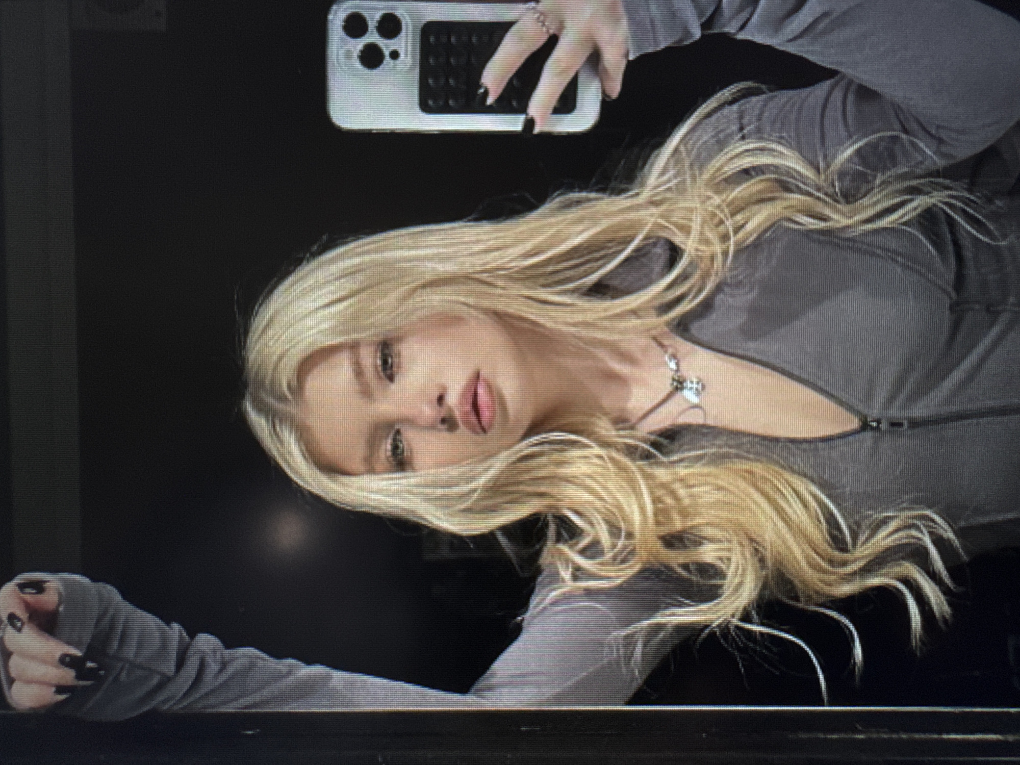 A young woman is depicted in what appears to be a selfie, lying down or leaning back against a dark, undifferentiated background. She has long, wavy blonde hair that is spread around her head and shoulders. Her eyes are light-colored, and her lips are slightly parted.
She is wearing a dark gray, long-sleeved top with a zippered neckline. A silver necklace with a small pendant is visible around her neck. Her fingernails are painted black on both visible hands. In her right hand, she holds a smartphone with a white case and a dark, multi-lens camera module, consistent with an iPhone. Her left hand and forearm are also visible in the lower left portion of the frame.
The setting appears to be indoors with subdued lighting. The image exhibits visible horizontal scan lines, suggesting it might be a photograph of a digital display rather than an original capture. No legible text is visible.