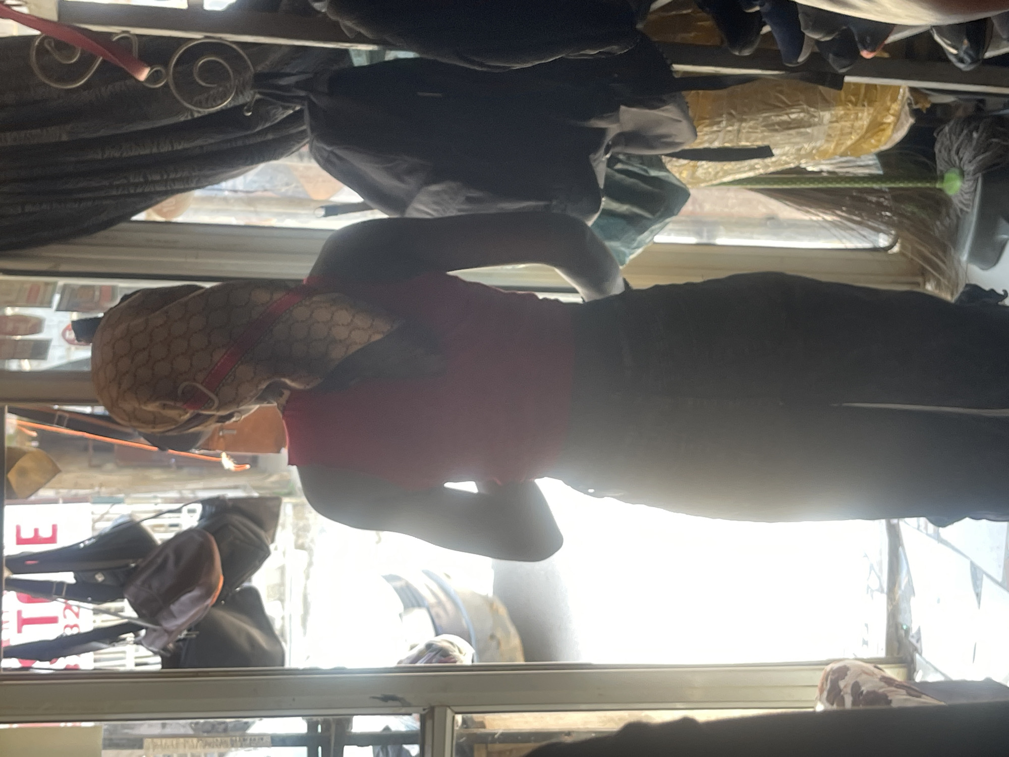 The image depicts the interior of a shop, likely a clothing or accessories store, in Abuja, Nigeria. The perspective is from behind a person wearing a pink sleeveless top and dark trousers, who is facing a doorway. The person is also wearing a patterned headscarf. Through the open doorway, bright daylight illuminates the exterior, revealing what appears to be a street scene with a vehicle. Inside the shop, various items are visible, including handbags hanging from the ceiling and a rack of clothing to the right. A broom and mop are positioned near the doorway. The overall environment suggests a commercial establishment in a Nigerian city.
