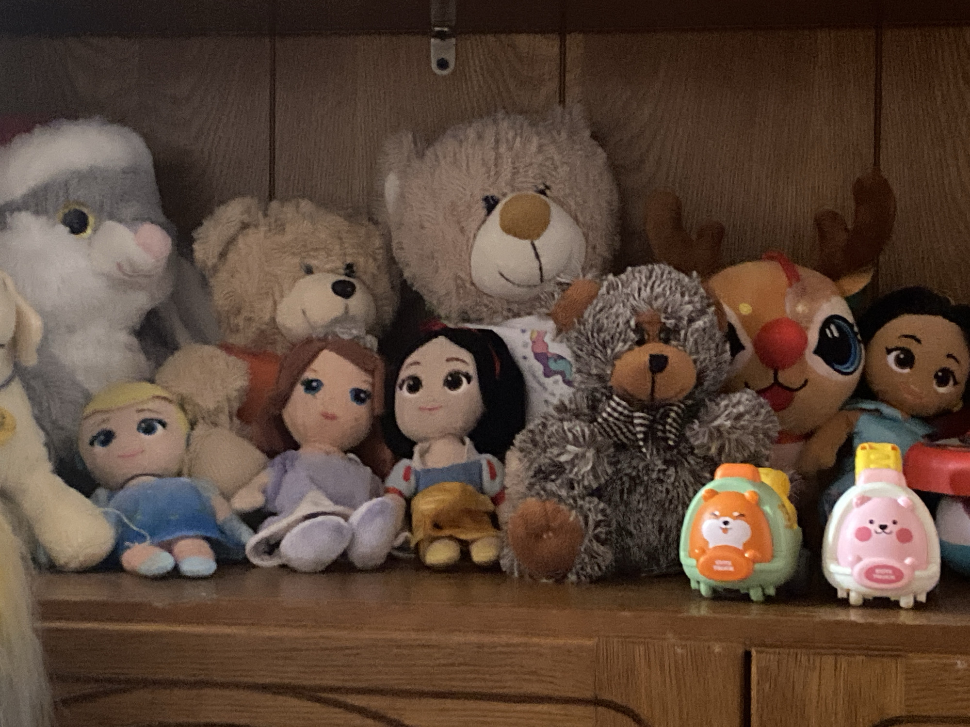 This is a medium shot of a collection of stuffed animals and two toy characters arranged on a wooden shelf. The stuffed animals include a large gray and white bunny, a tan teddy bear, a larger beige teddy bear, and a small fluffy gray and black teddy bear wearing a bowtie. Among the stuffed animals are two dolls, one dressed in a blue dress and the other in a purple dress. A doll resembling Snow White is also present, wearing her iconic dress. A stuffed reindeer with antlers is visible. In the foreground, two small toy characters with animal faces, one orange and one pink, are positioned on the shelf. The shelf is made of wood and appears to be part of a larger wooden cabinet or furniture piece. The background is a plain wooden surface, suggesting the shelf is inside a room or cabinet. The lighting is diffused and suggests an indoor environment. The scene is static, with no visible actions or interactions occurring.