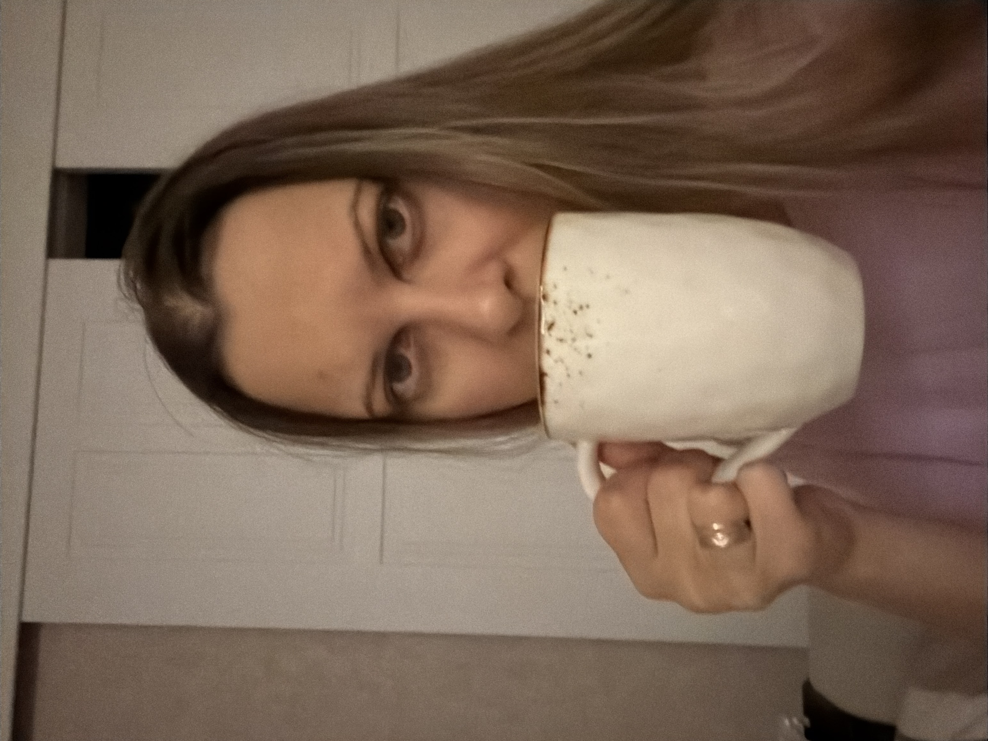 This portrait-style image, originally rotated 90 degrees counter-clockwise, depicts a woman indoors in Izhevsk, Russia. She has long, light brown hair and is looking directly at the viewer with a neutral gaze. Her right hand, wearing a gold-colored ring on the ring finger, holds a white mug with dark speckles to her mouth, suggesting she is drinking from it. The background features a light-colored wall or door, part of which has a dark rectangular insert. The image appears somewhat grainy. There is no visible text.