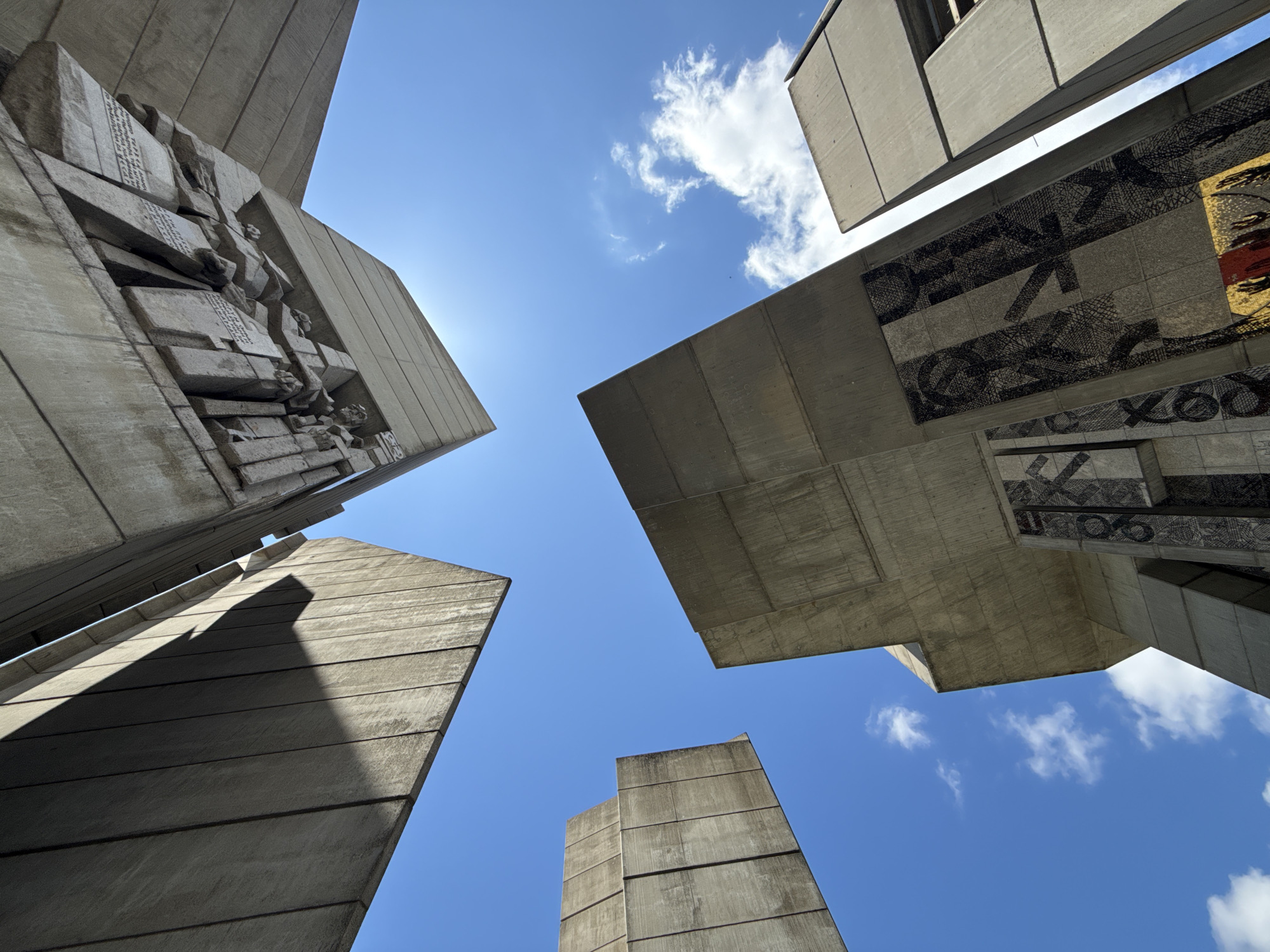 The image is a low-angle, upward view of the Monument to the 1300 Years of Bulgaria in Shumen, Bulgaria. The monument's imposing concrete structures dominate the frame against a clear blue sky with sparse white clouds. Sunlight casts sharp shadows on the textured concrete surfaces, highlighting geometric shapes and the brutalist architectural style.
In the upper part of the image, a large concrete slab extends downwards, featuring a carved relief with figures and what appear to be inscriptions. Below this, another massive concrete structure rises, adorned with mosaic panels. These mosaics display intricate patterns in black, white, and gold, with some sections featuring stylized lettering or symbols. To the right, the edge of another tall concrete pillar is visible.
The composition emphasizes the scale and monumental nature of the architecture, with the sky acting as a backdrop that contrasts with the solid, gray materials of the monument. There are no people or animals visible in the scene. The environment is clear and bright, suggesting a sunny day.