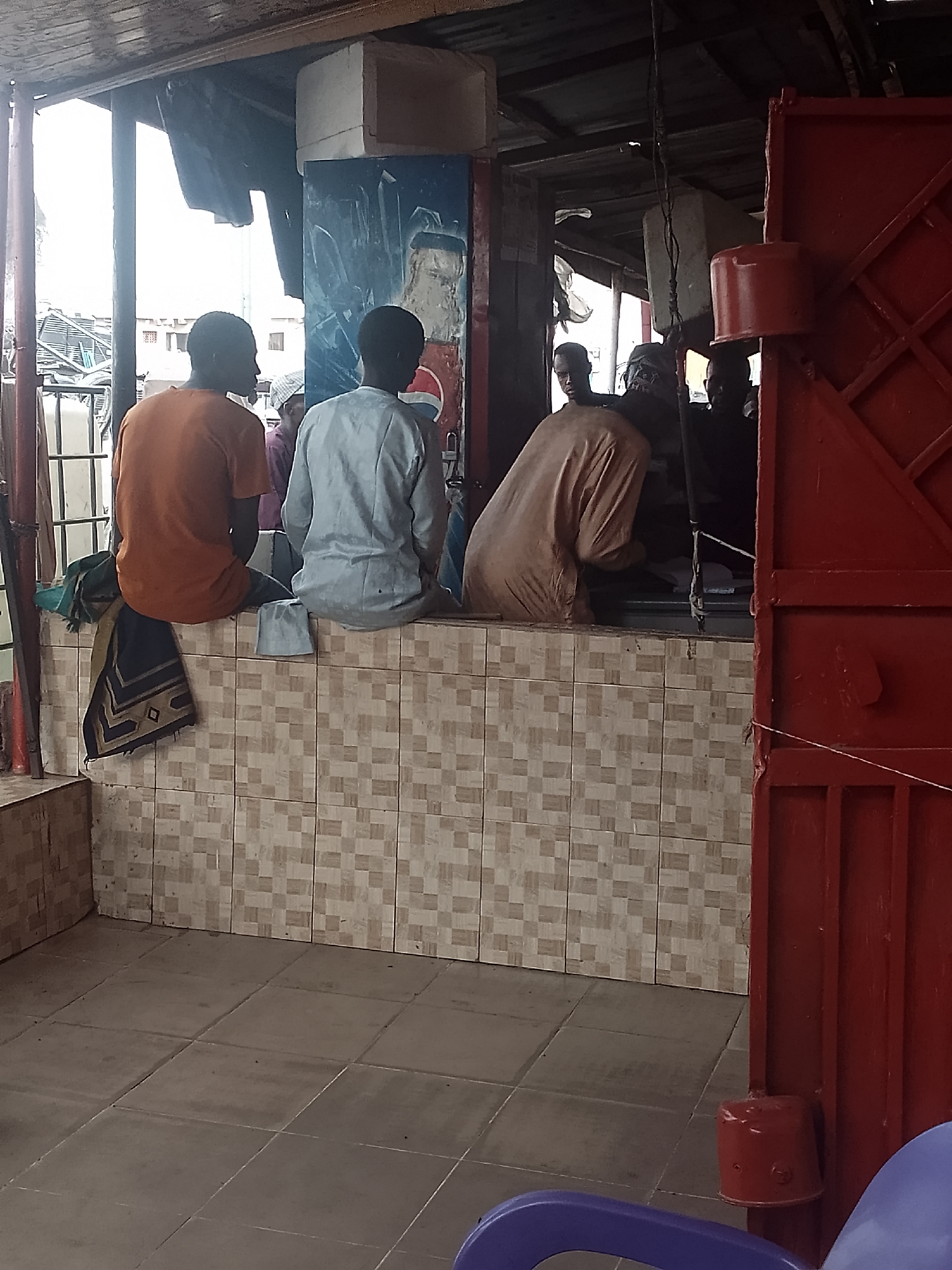 In Pantame, Nigeria, a group of men are seated on a tiled structure. The men are wearing traditional clothing. A red gate is visible on the right. The location appears to be an outdoor space with a covered roof. There is a beverage advertisement on the left.