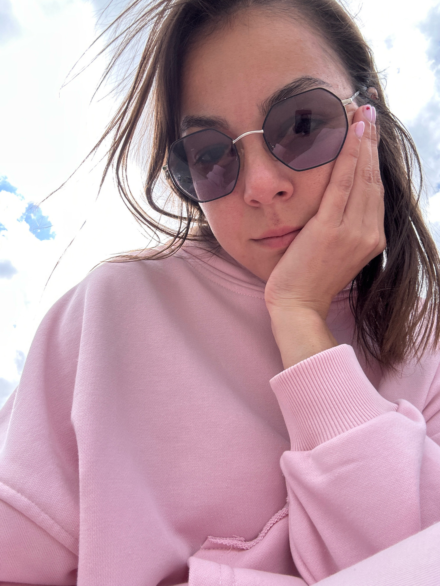 The image is a close-up, low-angle selfie of a young woman with dark hair and fair skin, wearing oversized, dark-tinted hexagonal sunglasses and a pale pink hoodie. She is resting her right cheek on the back of her right hand, with her fingers extended up towards her temple. Her left hand is not visible. Her fingernails are painted a light pink. The background consists of a bright, overcast sky with patches of blue. The woman is looking downwards, away from the camera, with a pensive expression. The lighting suggests it is daytime. There is no indication of the location being in Gyumri, Armenia.