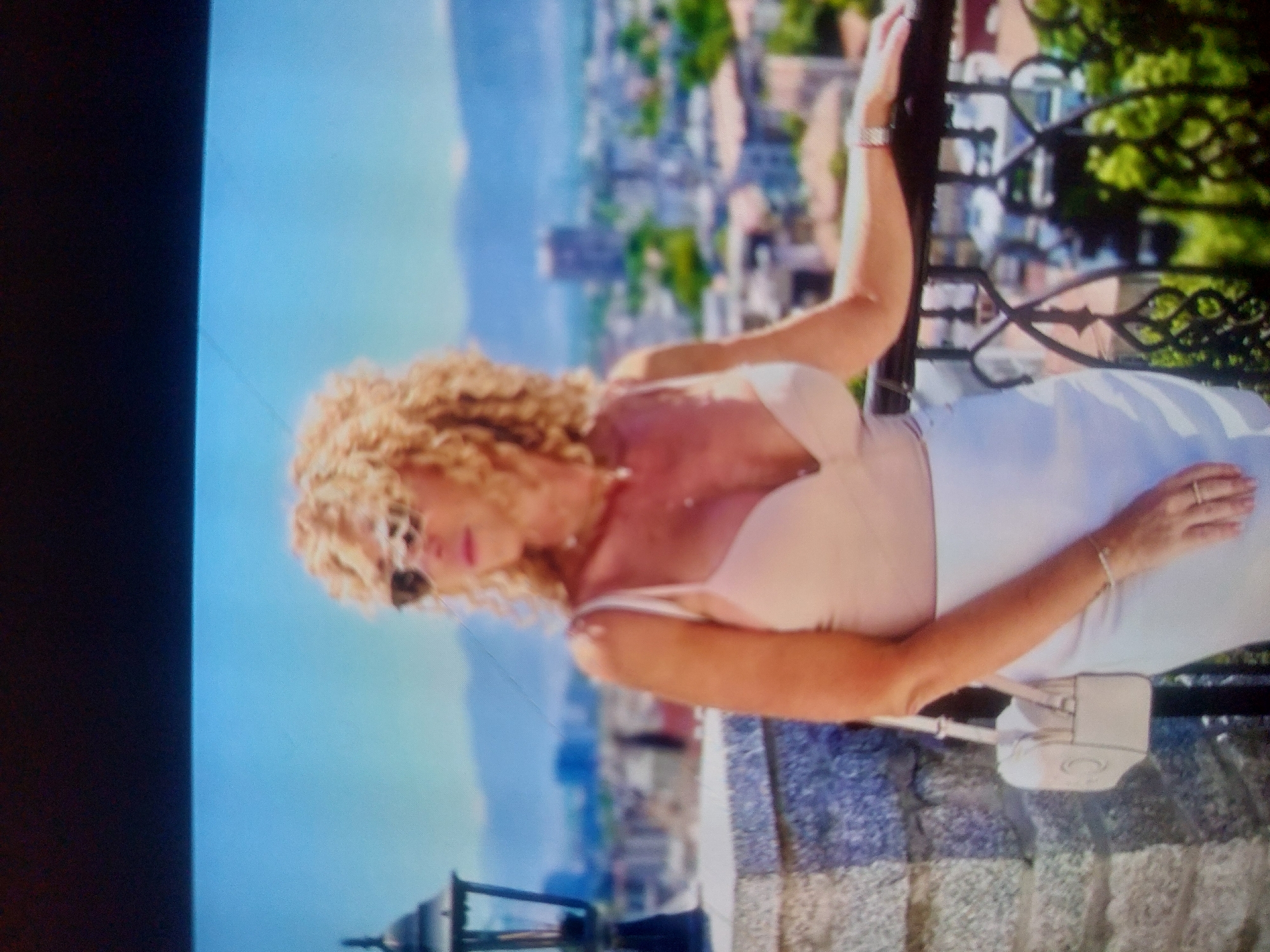 A medium shot of a fair-skinned woman with blonde curly hair wearing sunglasses, a low-cut top, and a white skirt, leaning against a metal railing on a stone balcony. She has a light-colored purse hanging from her shoulder. In the background is a cityscape, with buildings, trees, and a mountain range, possibly in Tsarevo, Bulgaria. A black object, possibly a light fixture, is in the lower left corner.