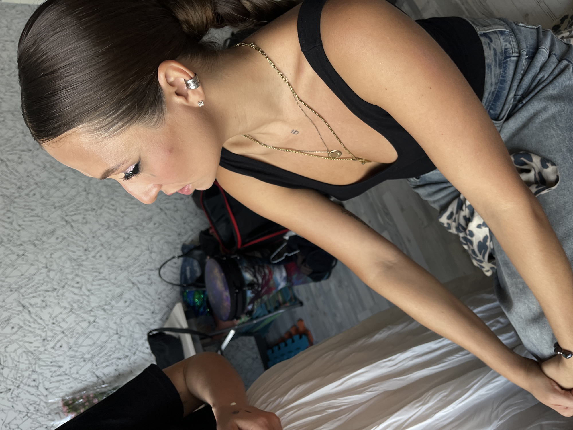 An unidentifiable young woman with dark hair pulled back in a bun, wearing a black tank top and blue jeans, is seated indoors. She is adorned with jewelry, including a silver ear cuff, a stud earring, and two gold necklaces. Her makeup includes glittery eyeshadow and winged eyeliner. Her arms are crossed in front of her, and her hands are clasped. To her left, another person, only their arm and shoulder are visible, wearing a black t-shirt and a long white skirt, is also seated. In the background, various objects are visible, including a bouquet of pink flowers, bags, and what appears to be a large drum-like object with colorful patterns. The wall behind them has a textured, gray pattern. The lighting suggests an indoor setting, possibly a room. There is no indication of the specific location within Moscow, Russia.