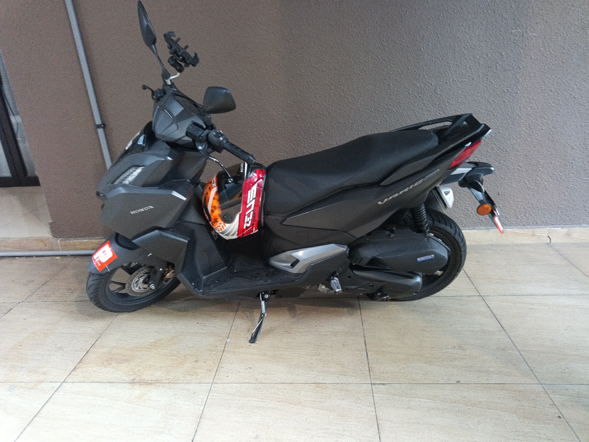 A black Honda motorcycle with a red and white sticker on the rear fender is parked on a tiled surface in front of a textured brown wall in Kuala Lumpur, Malaysia. The motorcycle is equipped with a phone mount on the handlebars, and a helmet with orange and white detailing is clipped to the side. The kickstand is deployed. The scene appears to be an outdoor area, possibly a residential or commercial property, given the wall and tiled flooring. No individuals are present in the image.