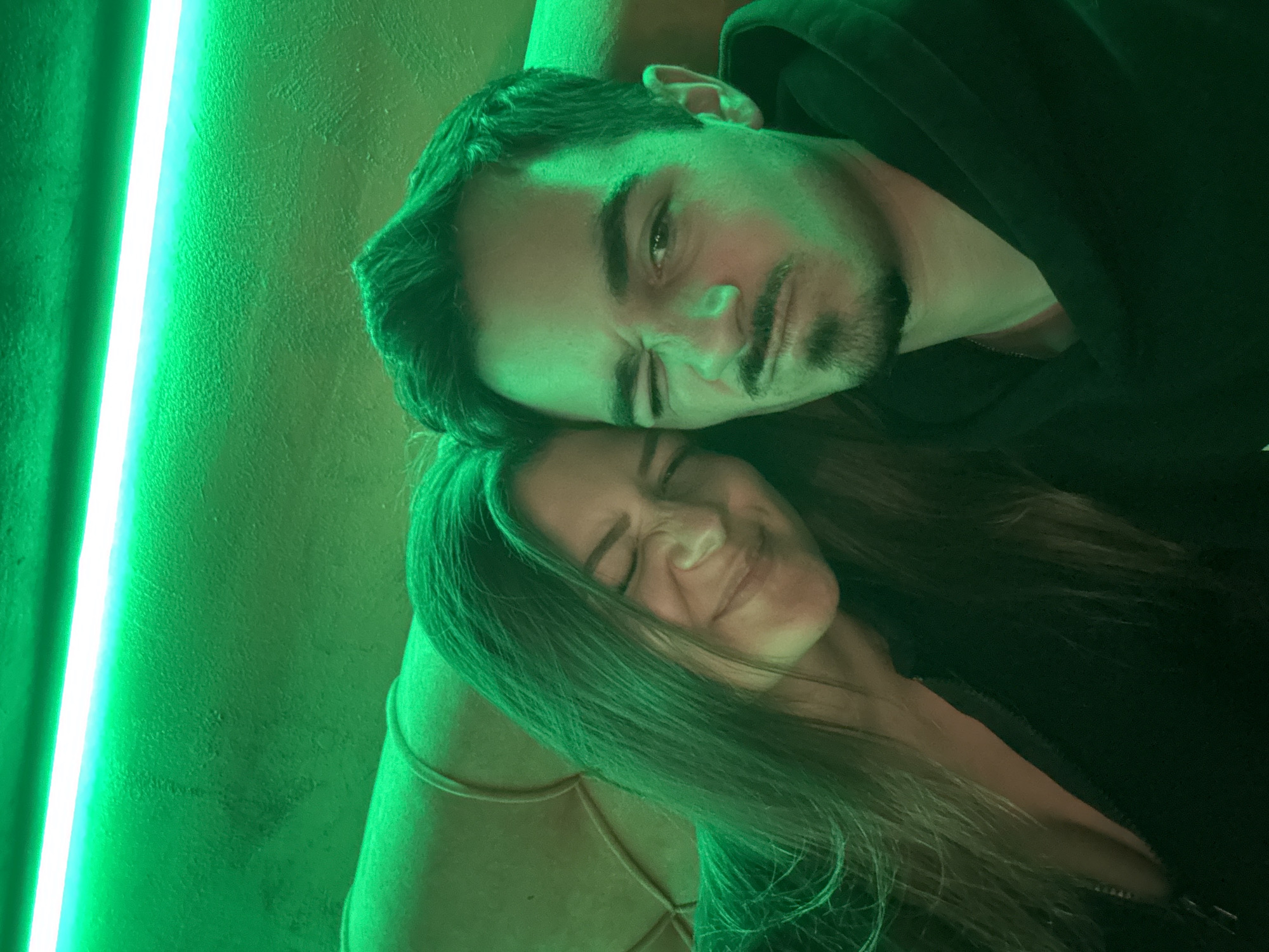 A photograph depicts a man and a woman posing closely together, bathed in a green-toned light. The man, with short dark hair and a beard, is wearing a black hooded garment. He is squinting his eyes, suggesting a playful expression. The woman, with long brown hair, is smiling with her eyes closed. Both individuals are wearing dark-colored clothing. To the left, a bright, linear green light source is visible, positioned vertically against a textured wall. No specific location details within Sofia, Bulgaria are discernible from the image.