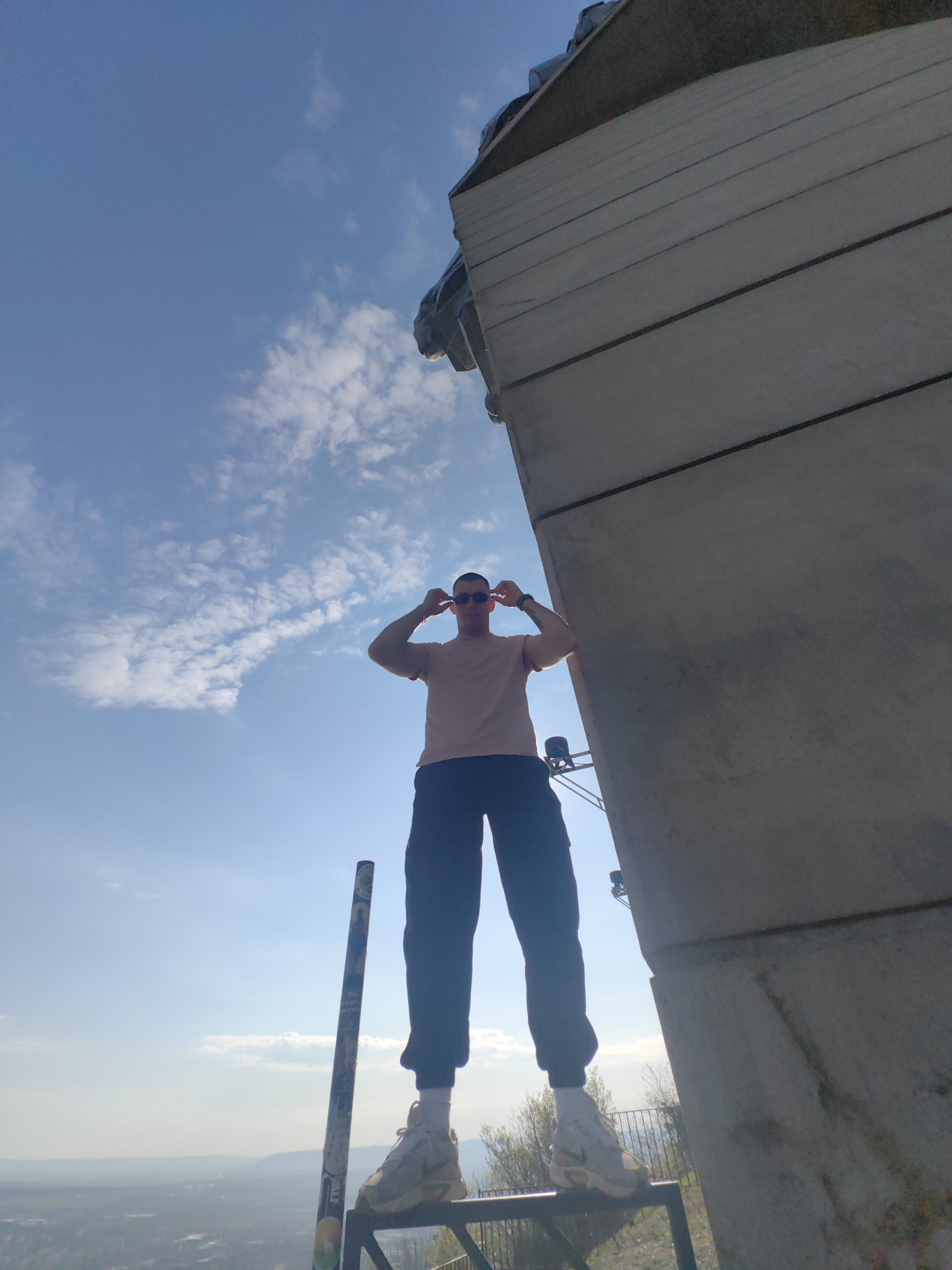 A low-angle, upward-facing shot depicts a man in sunglasses standing near a monument, likely in Shumen, Bulgaria. The man wears a pink short-sleeved shirt, dark-colored pants, and white sneakers. He is holding his sunglasses with both hands, seemingly adjusting them. He stands on a dark gray metal structure which is attached to a concrete monument. The monument is positioned to the right of the man and fills a significant portion of the frame with gray, layered concrete panels. A blue sky filled with light, scattered clouds provides the backdrop. In the distance, a cityscape or landscape is visible. A pole with stickers or markings stands next to the man and to the left.