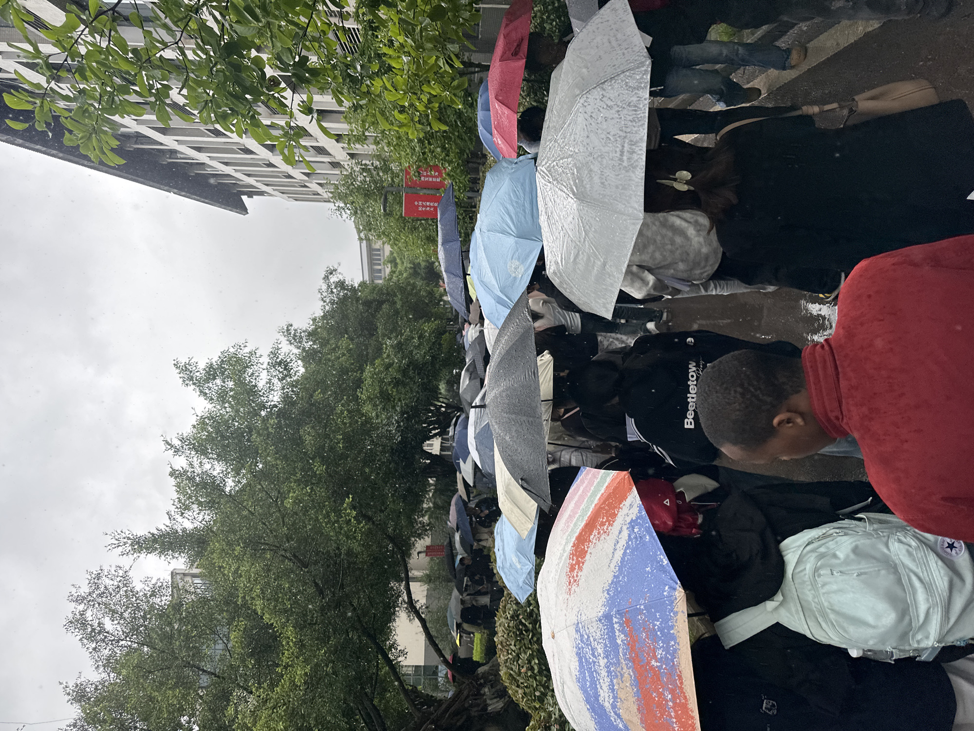 The image captures a crowded outdoor scene in Jiangjin, China, during a rainy day. Numerous people are gathered, forming a long line or queue, with most sheltering under a variety of umbrellas. The scene is shot from behind the line, looking forward.
In the foreground and midground, a dense group of individuals stands close together. Most of their bodies are obscured by the many umbrellas they hold, which range in color from solid red, blue, grey, and white to a vibrant multi-colored one in the immediate foreground, displaying streaks of blue, red, orange, and white. Water droplets are visible on the surfaces of the umbrellas, indicating active rainfall. People are dressed in casual attire; one person wears a red top and another a black shirt with "Beetletow" visible text, while a light blue fanny pack with a Converse logo is also seen. The ground beneath them appears wet and reflects some light.
In the background, tall, multi-story buildings with repetitive window patterns are visible on the left, partially obscured by lush green trees with dense foliage. A red rectangular sign with white Chinese characters is mounted on a pole near the buildings and trees. The text reads, from top to bottom: "禁止停车" (jìnzhǐ tíngchē - No Parking), "消防通道" (xiāofáng tōngdào - Fire Lane), and "违者必究" (wéizhě bìjiū - Violators will be prosecuted).
The weather is overcast, with a bright, cloudy sky dominating the upper part of the background. The overall impression is one of a busy, damp day, likely in a university or urban setting, where people are patiently waiting in line despite the rain.
