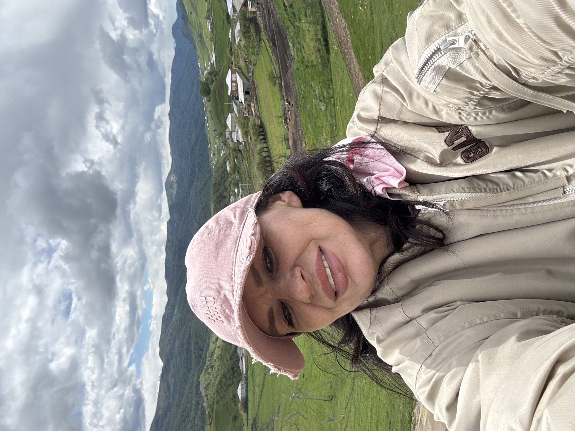 A woman in a beige jacket and pink cap smiles for a selfie. The background shows a green, mountainous landscape with small buildings clustered together. The sky is cloudy. The photo appears to be taken in Верхняя Мара, Russia.