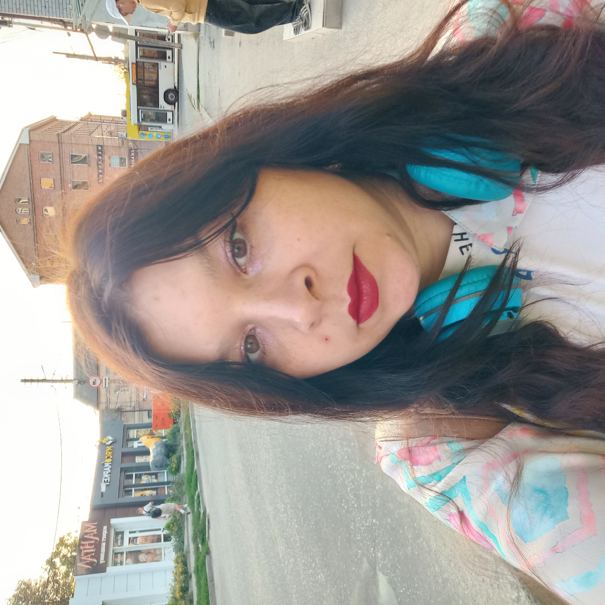 The image is a selfie taken outdoors on a street, likely in the city of Kropyvnytskyi, Ukraine. The foreground is dominated by the face of a woman with long, dark brown hair, wearing blue headphones around her neck and a colorful patterned shirt. She has bright makeup on her eyes and lips. In the midground, a street with a sidewalk is visible. On the sidewalk, a man in a yellow hoodie and dark pants is standing, looking at his phone. Behind him, a white bus with illuminated signs is passing by. Further in the background, there are buildings, one of which is a multi-story brick structure with visible windows, and another appears to be a commercial building with signage. Trees and utility poles are also part of the environment. The lighting suggests it is either morning or late afternoon.