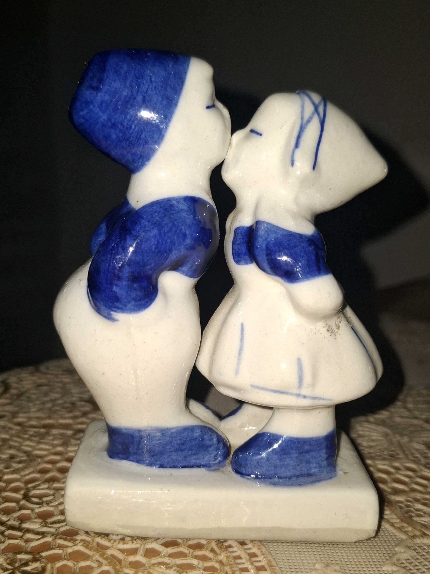A small, white porcelain figurine depicts two children, a boy and a girl, sharing a kiss. The boy, on the left, wears a blue cap and blue accents on his body. The girl, on the right, wears a white dress with blue accents and blue lines on her headpiece. They stand on a white rectangular base. The figurine is placed on a beige lace tablecloth. The background is dark and out of focus, suggesting an indoor setting. The location of this image in Almaty, Kazakhstan, cannot be determined from the visual information provided.