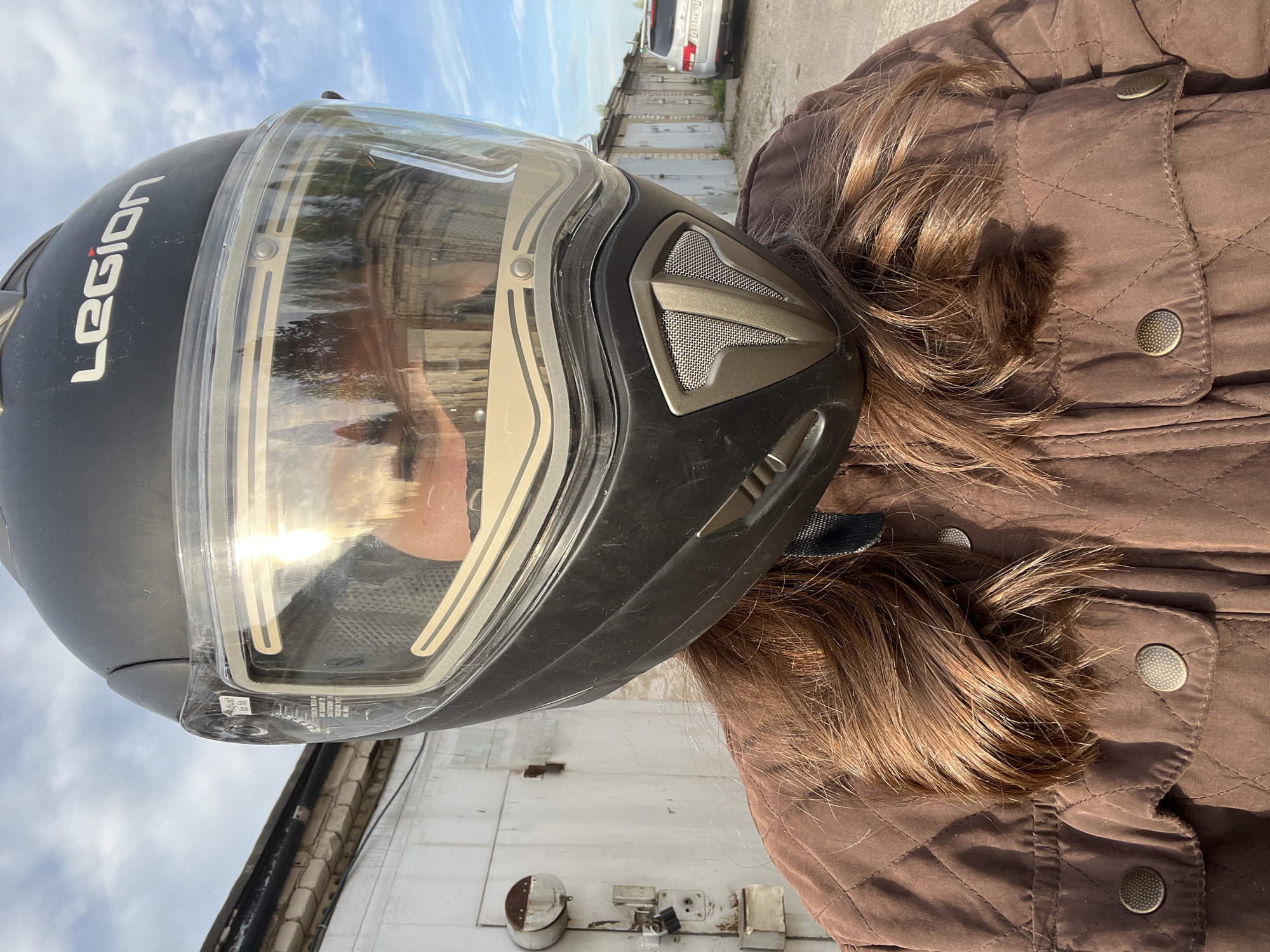 A person wearing a black motorcycle helmet labeled "LEGION" and a brown quilted jacket is the primary subject. The helmet has a clear visor reflecting sunlight. The person's hair is brown. The background includes a white building with a drainpipe, possibly a garage, and a vehicle partially visible in the upper center of the frame. The location is identified as Chaikovsky, Russia.