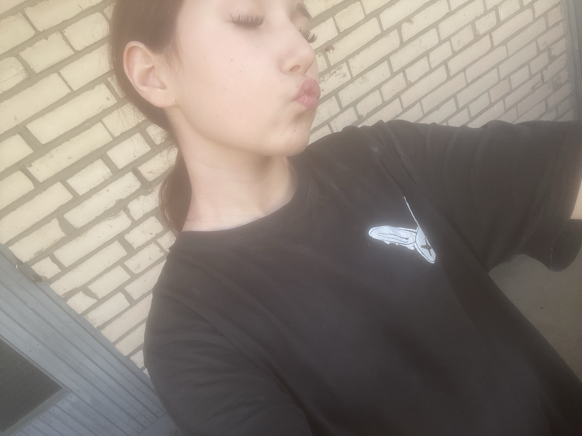A medium close-up photograph features a young person posing with a playful expression, appearing to blow a kiss. The subject is seen in profile, facing towards the left side of the frame, with their eyes closed or cast downwards and lips pursed. Long, prominent eyelashes are visible. Their hair, dark brown, is pulled back into a low ponytail, and part of their right ear is visible.
The person is wearing a dark, likely black, short-sleeved t-shirt. On the left chest area of the t-shirt, there is a distinct, stylized white graphic resembling a shoe or boot, possibly a brand logo.
The background consists primarily of a light-colored brick wall with darker mortar lines, suggesting an outdoor setting or an indoor space with an exposed brick wall. Below and to the left, another section of the background shows horizontal grey slats, which could be part of a window frame, siding, or another architectural element. The lighting is bright and even, indicative of daytime. The overall mood of the image is lighthearted and candid. The photograph is understood to have been taken in Saint Petersburg, Russia. No discernible text is visible other than the graphic logo.