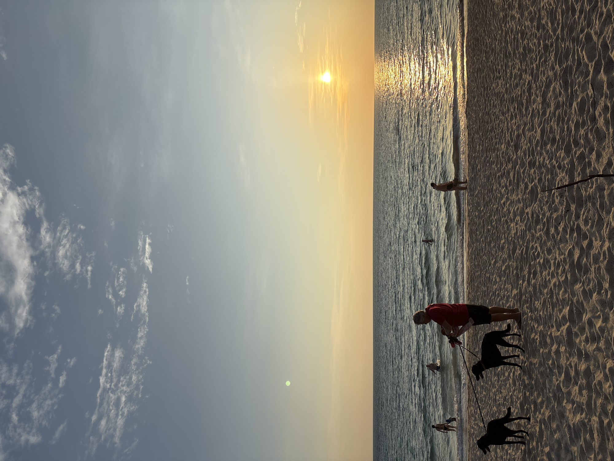 A person with two black dogs is walking on the sand at a beach in Choeng Thale, Thailand. The sun is setting over the ocean. Other people are in the water and on the beach. The sky has clouds.