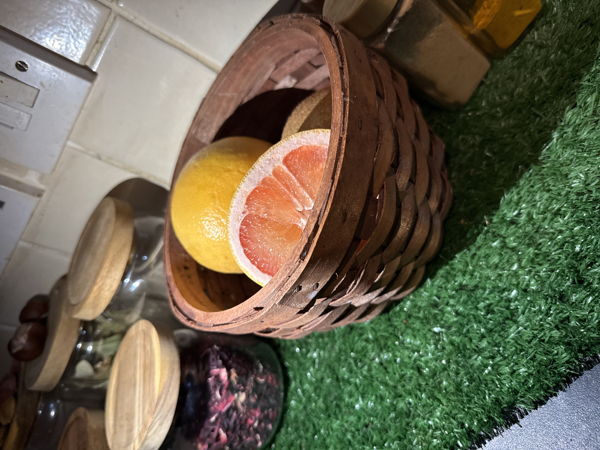 An indoor, close-up photograph displays a still life arrangement, taken from a slightly elevated, diagonal perspective. The central subject is a rustic, woven brown basket containing two grapefruits. One grapefruit is whole and yellow, while the other is cut in half, revealing its vibrant pink and segmented interior.
To the right and below the basket, a patch of bright green artificial grass, resembling turf, covers the surface, transitioning to a dark, possibly grey or black, surface in the bottom right corner. In the background, to the left of the basket, there is a light-colored tiled wall with visible grout lines and an electrical outlet or light switch mounted on it. Below this, partially obscured, are several clear glass jars with wooden lids. One jar clearly holds a reddish-purple substance, possibly dried berries or tea leaves. Above and behind the basket, a glass container with a yellow liquid, likely honey or oil, is visible with a wooden lid.
The scene depicts a domestic setting, possibly a kitchen or a decorative display area. The electrical outlet type is consistent with those found in the United Kingdom, aligning with the provided location context of Great Yarmouth. No specific activity is occurring, and there is no visible text. The lighting appears to be artificial, casting some soft shadows.