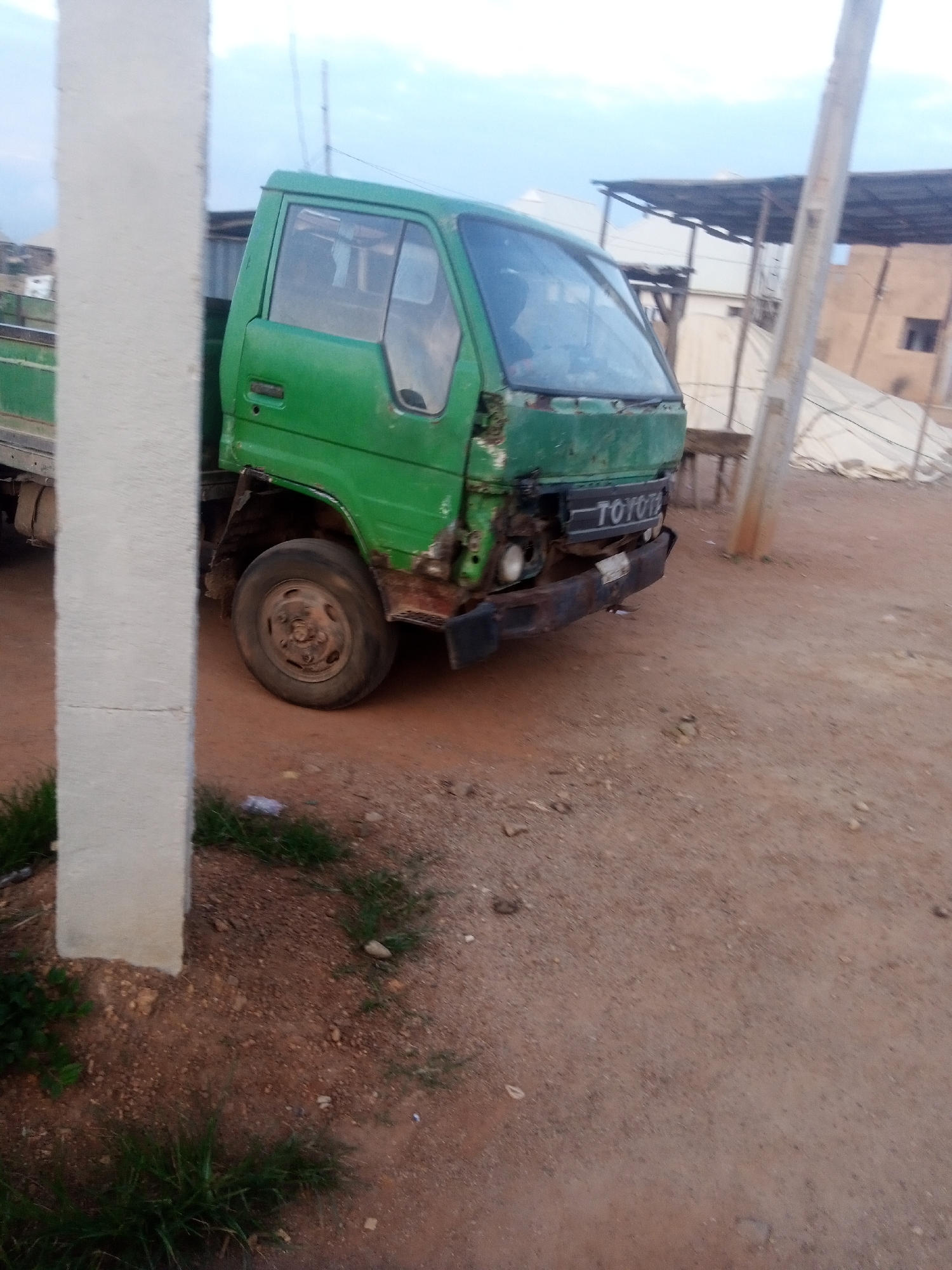 The image shows a damaged green Toyota truck parked on a dirt road. The truck is the main subject, with its front bumper and grill clearly damaged. The "TOYOTA" logo is visible on the front. The truck appears to be old and weathered, with rust visible on its body.
The scene is an outdoor setting, likely a yard or a roadside. There is some sparse vegetation on the ground and a concrete pillar on the left side of the image. In the background, there are some structures, including what appears to be a covered area with corrugated metal roofing, and other buildings. A utility pole is also visible to the right of the truck.
The lighting suggests it might be late afternoon or early morning, with soft light illuminating the scene. The sky is partly cloudy. There are no people visible in the image. The overall impression is one of a functional, but aging, vehicle in an unpaved, possibly rural or industrial, area.