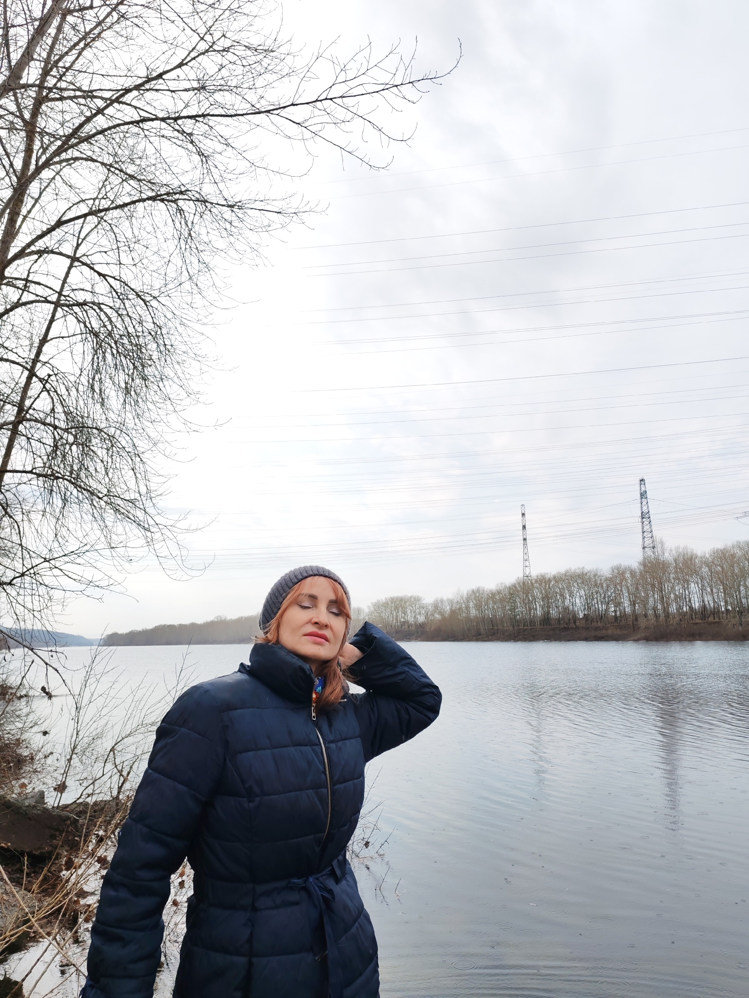 A woman wearing a dark blue puffy coat and a gray beanie stands on the shore of a body of water. Her eyes are closed and her right hand is raised to her head. Behind her is a barren tree on the left, the water of a lake in the center, and power lines stretching across the gray sky in the background. The opposite shore of the lake has trees. This image was taken in the city of Chaikovsky, Russia.