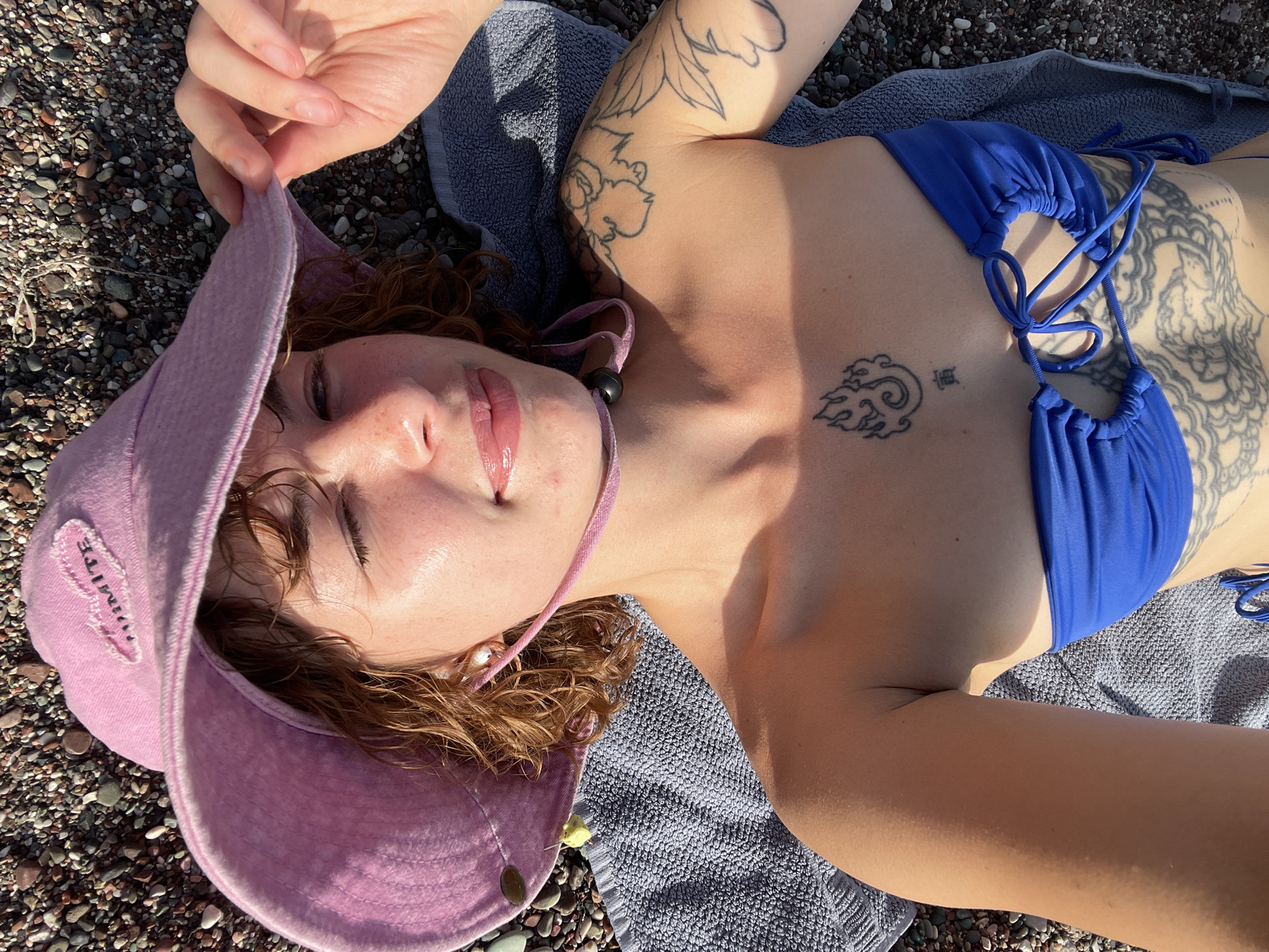 An overhead, close-up shot captures a young adult female relaxing on a pebble beach in Rafailovići, Montenegro. She is lying on her back on a gray towel, surrounded by small, dark-colored pebbles.
The woman has fair skin, highlighted by direct sunlight, and reddish-brown, curly hair. She is wearing a bright blue triangle-style bikini top with crisscrossing ties on the front. A light pink bucket hat is placed on her head, with one hand gently holding its brim. Her eyes appear partially closed, and her lips are visibly glossy, reflecting the sun. A small black bead or charm hangs on a string near her neck.
Several tattoos adorn her body: a floral design on her left upper arm, a stylized cloud or wave pattern alongside a character on her chest/sternum, and a large, intricate design, possibly a dragon, on her right side, partially visible beneath her bikini top. The hat features embroidered text, clearly displaying "TRUMITE".
The scene is brightly lit by strong, direct sunlight, indicated by the sharp shadows and the sheen on her skin and lips, suggesting a clear, sunny day. The overall impression is one of peaceful relaxation, as she appears to be sunbathing and enjoying the coastal environment.