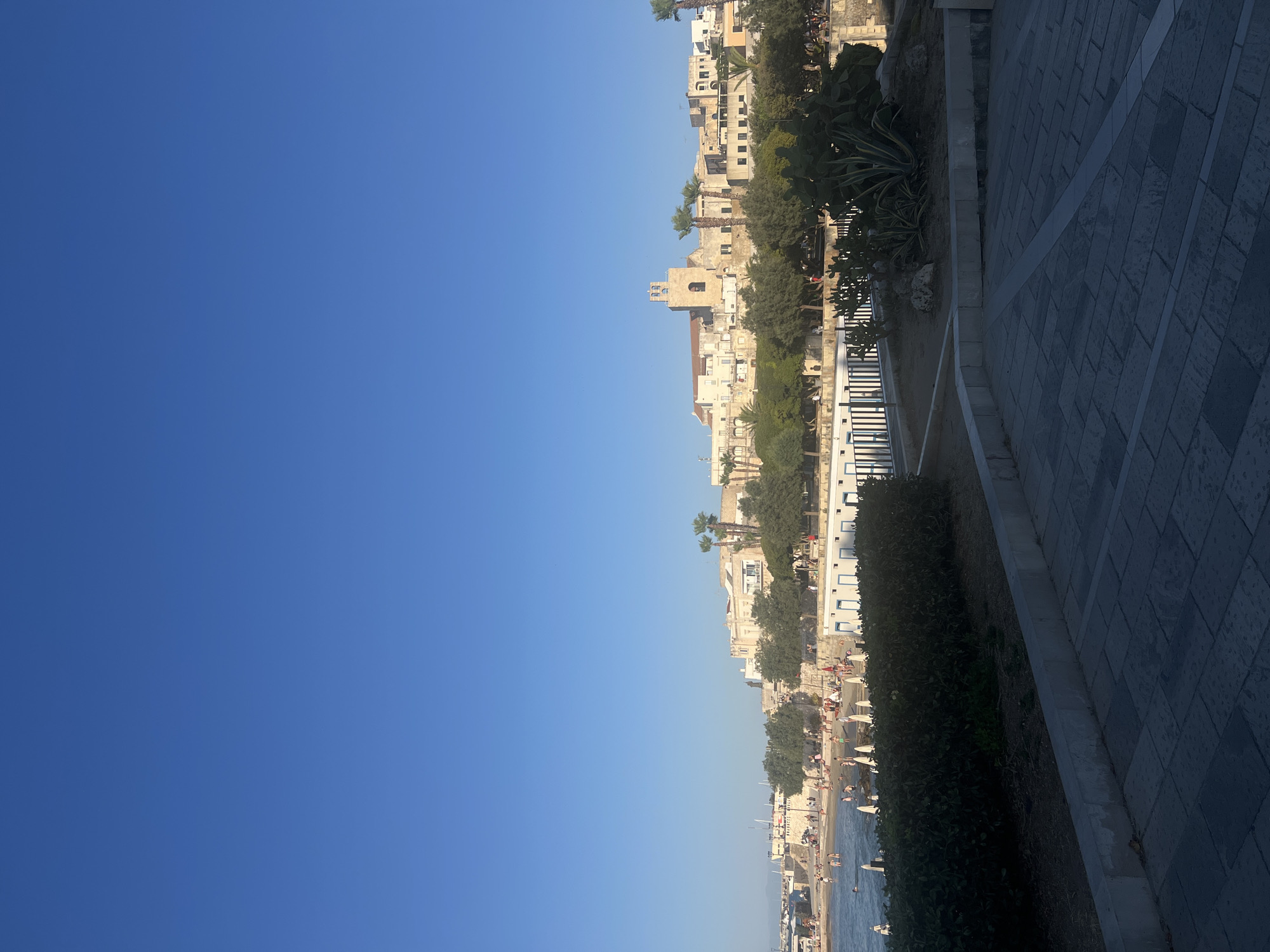 This is an outdoor, medium-shot, eye-level, vertical, still photograph of the waterfront in Otranto, Italy, on a clear, sunny day. The sky is a vibrant, cloudless blue, taking up the top two-thirds of the frame. The bottom third features the Otranto waterfront, with a beach, buildings, trees, and a paved area in the foreground.
The beach is lined with white beach umbrellas and has numerous people enjoying the water and sand. The water is calm and blue. Behind the beach, there is a row of white buildings with blue trim, possibly changing rooms or concessions. A line of trees, including palm trees, separates the beach area from the buildings of the town. The town itself is composed of light-colored, multi-story buildings, with a prominent bell tower of a church visible.
In the foreground, there is a paved walkway made of gray stone tiles, with some concrete curbing. Lush green bushes and some succulents, including a large agave plant, are visible in the middle ground, adding to the Mediterranean feel of the scene. The lighting suggests it is late afternoon, with long shadows cast on the paved area.