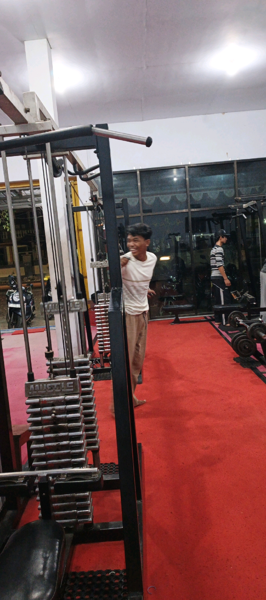 A young man with dark hair and a striped t-shirt is laughing in a gym in Kasiyan Timur, Indonesia. He is standing next to a weight machine. The gym has a red floor and a large window in the background. Another man in a striped shirt and a cap is visible in the background, looking down. There are motorcycles parked outside the gym, visible through the window.